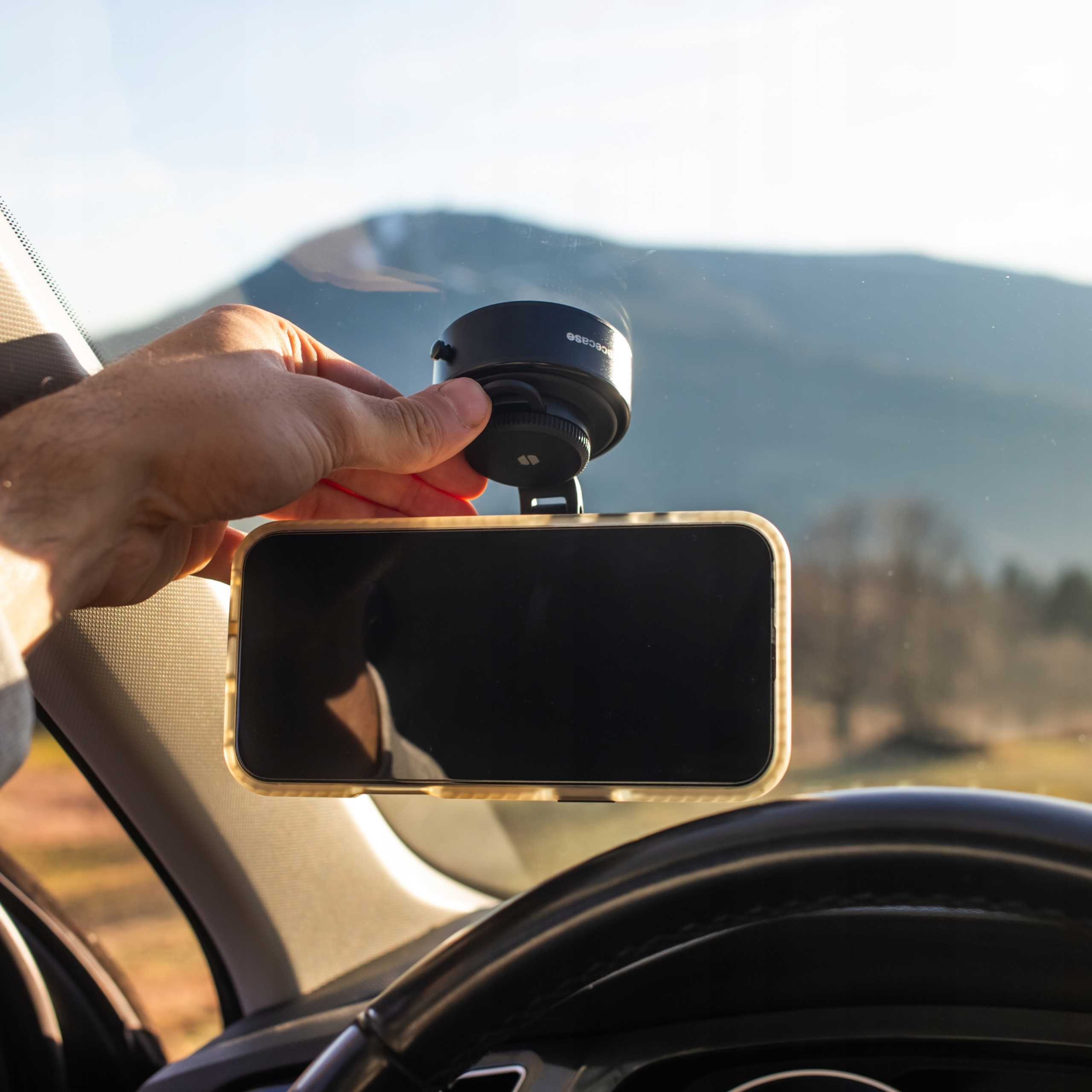 Dłoń trzyma telefon zamontowany na przedniej szybie samochodu. W tle widoczne są góry.
