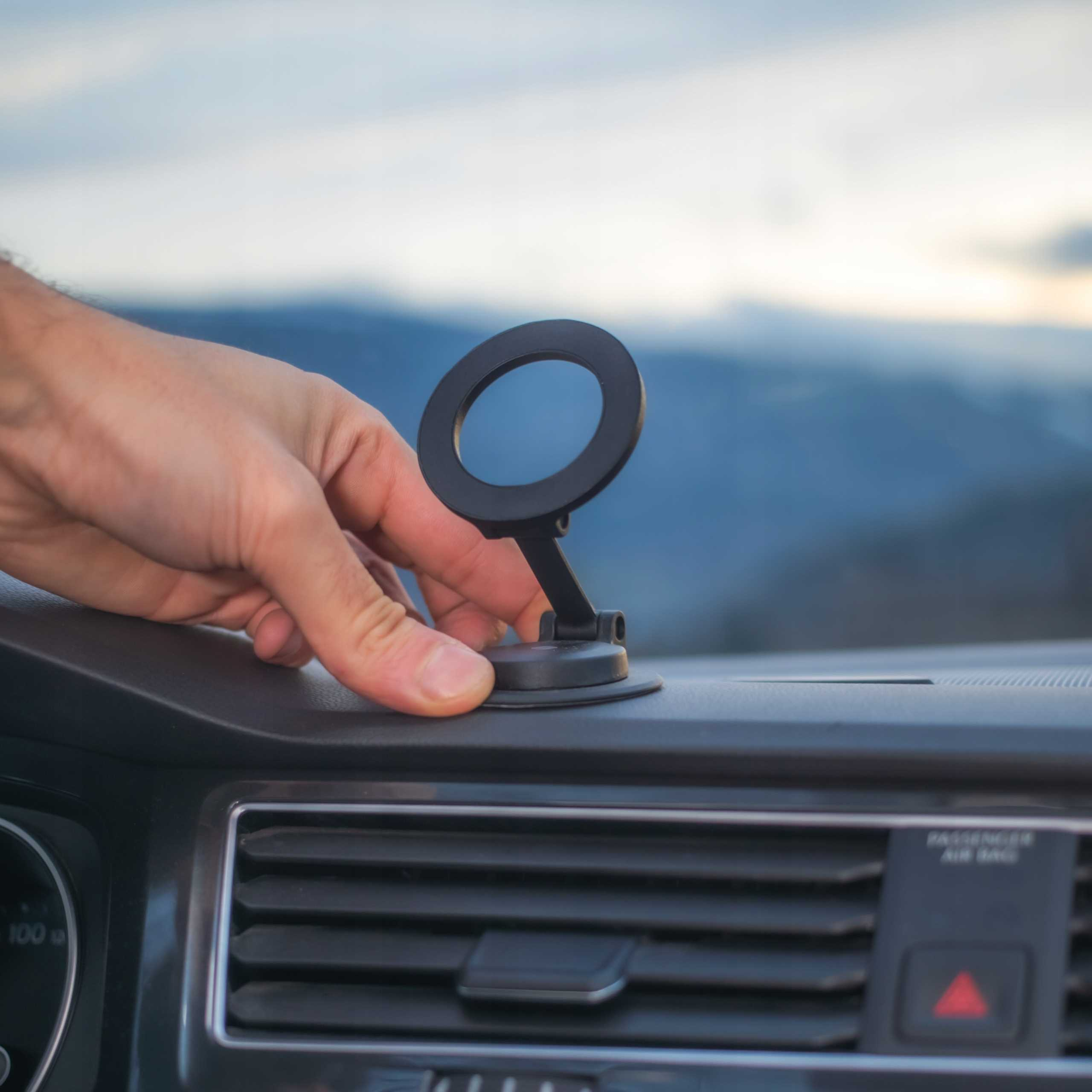 Ręka umieszcza czarny uchwyt na telefonie na desce rozdzielczej samochodu. W tle góry i niebo. Widoczne wnętrze samochodu.