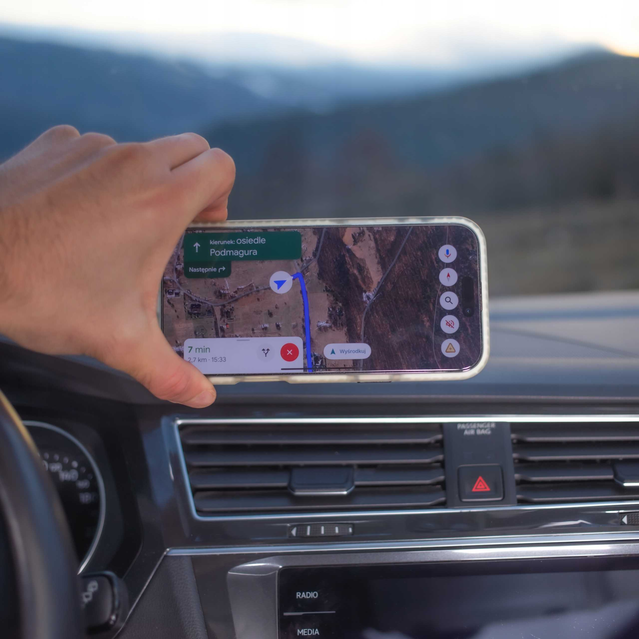 Ręka trzyma telefon z mapą w samochodzie. W tle widać góry przez przednią szybę.