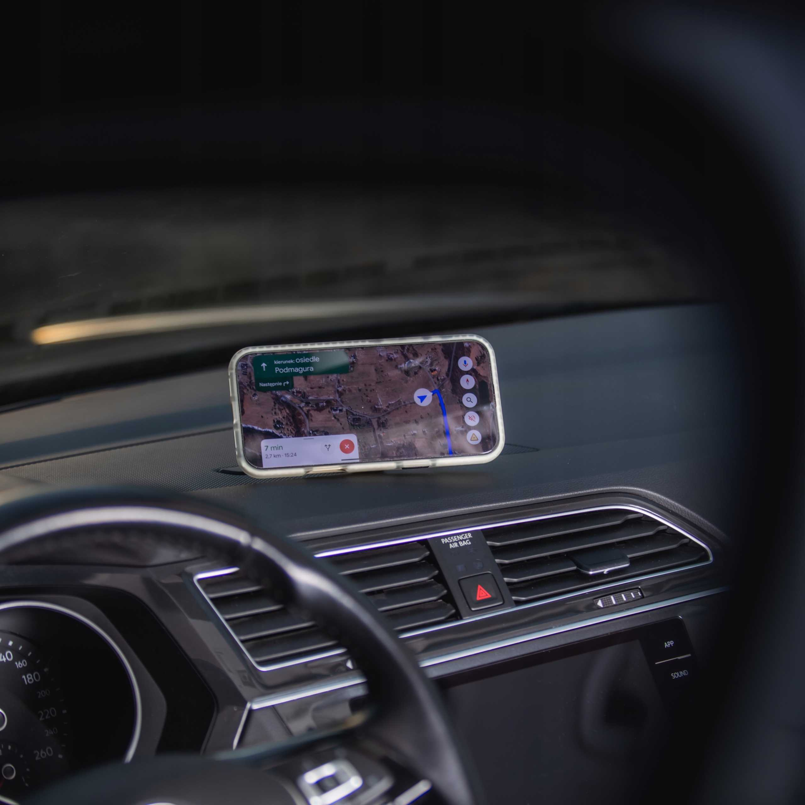 Telefon z nawigacją leży na desce rozdzielczej samochodu. Widoczne są kierownica i kratki wentylacyjne.