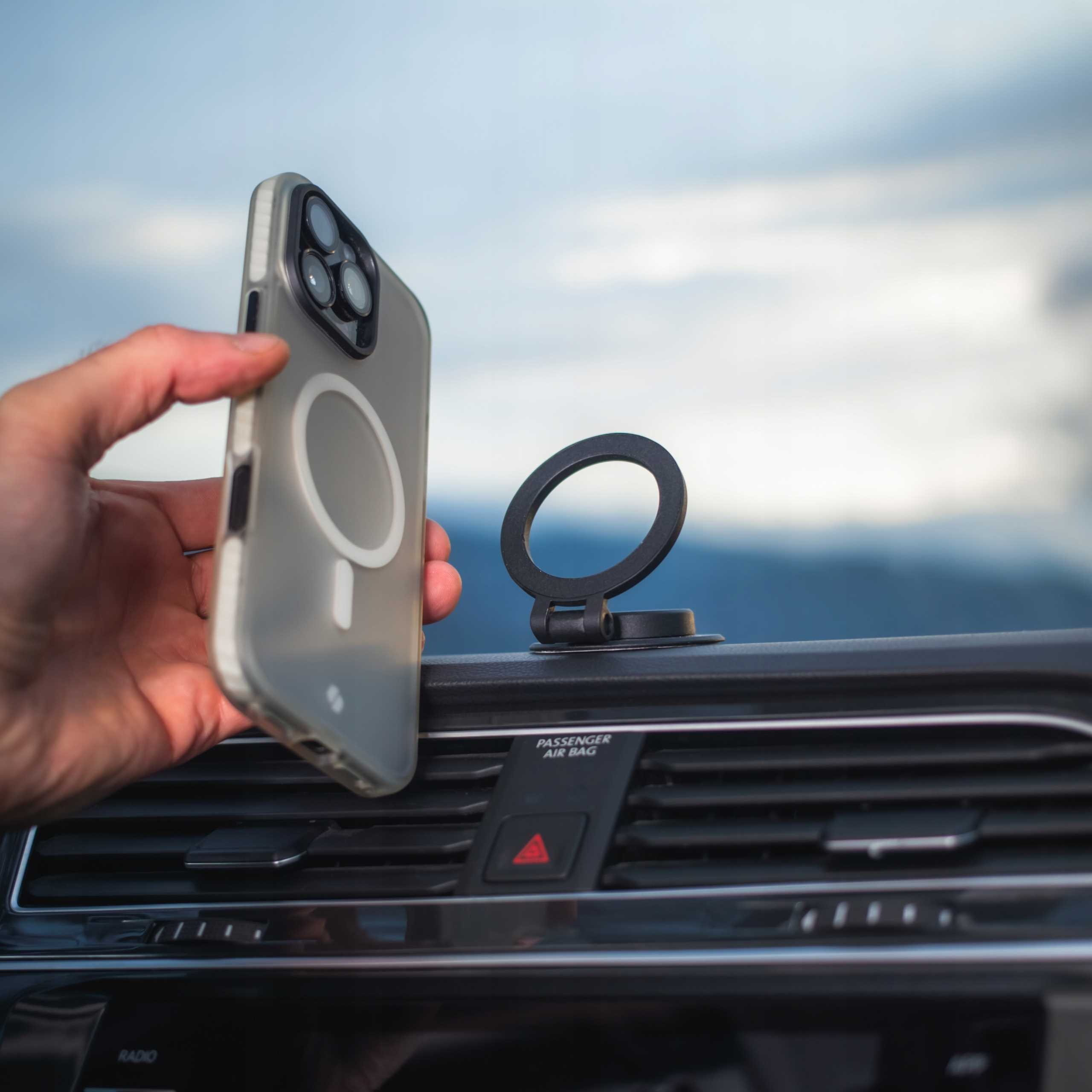 Ręka trzyma telefon w etui obok kratki wentylacyjnej samochodu. Czarny uchwyt na telefon jest zamontowany.