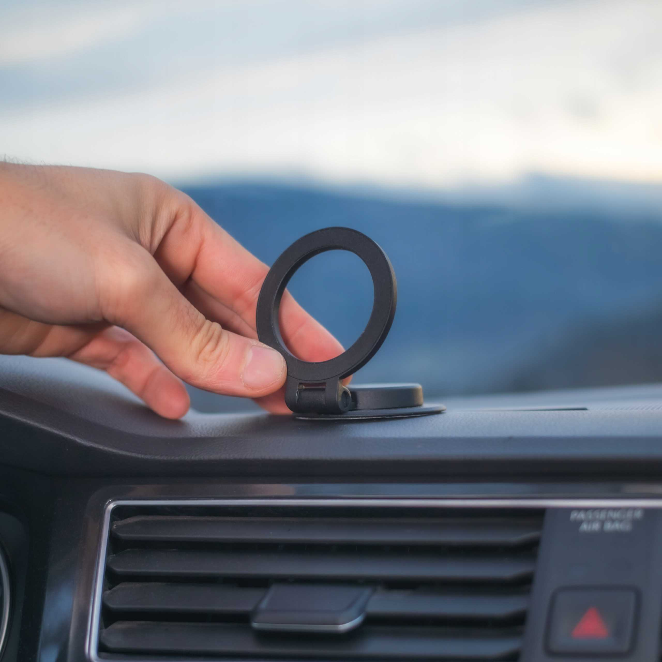 Ręka umieszcza czarny uchwyt na telefonie na desce rozdzielczej samochodu. Tło pokazuje góry i niebo.