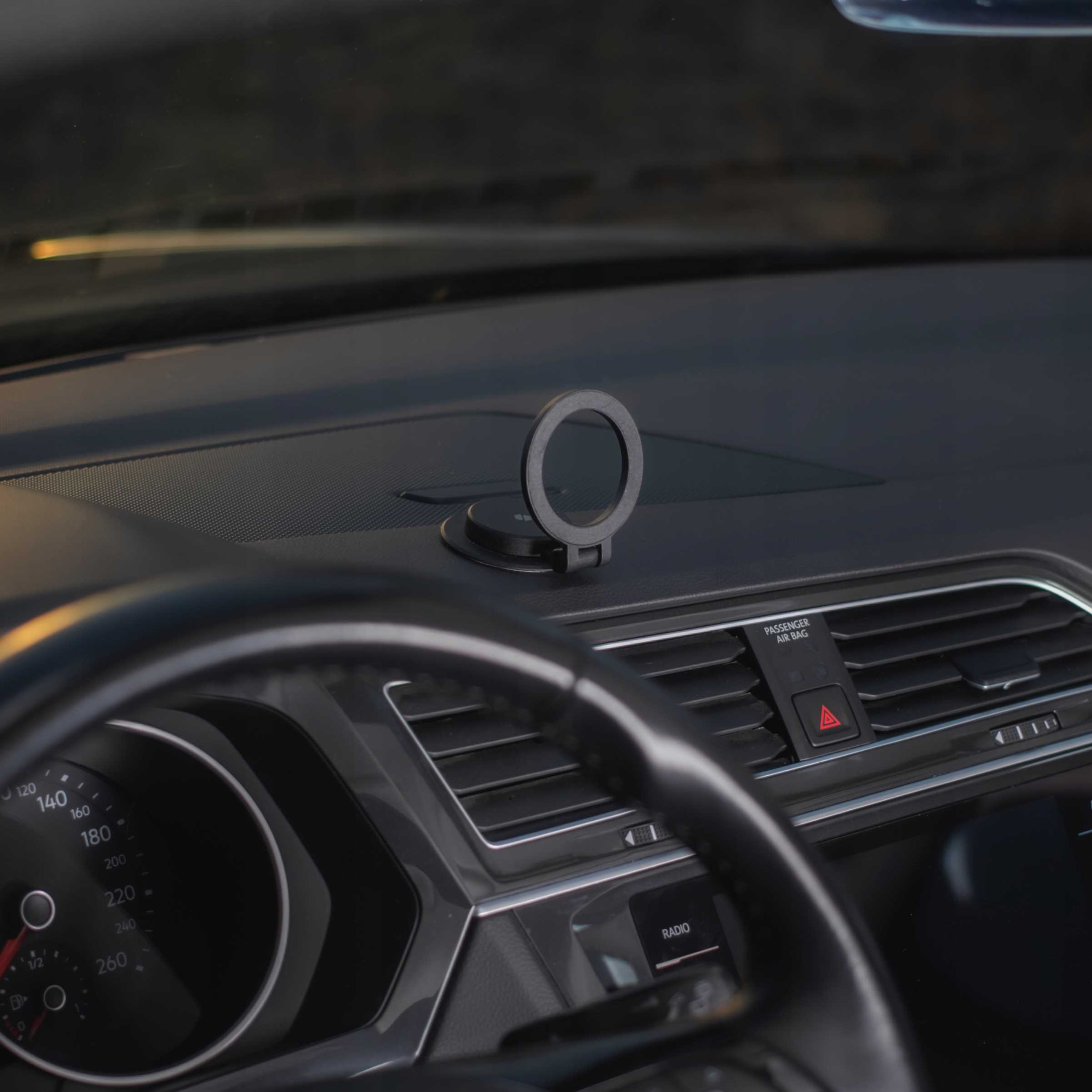Wnętrze samochodu. Deska rozdzielcza z okrągłym uchwytem na telefon. Kierownica z przodu, widoczny prędkościomierz.