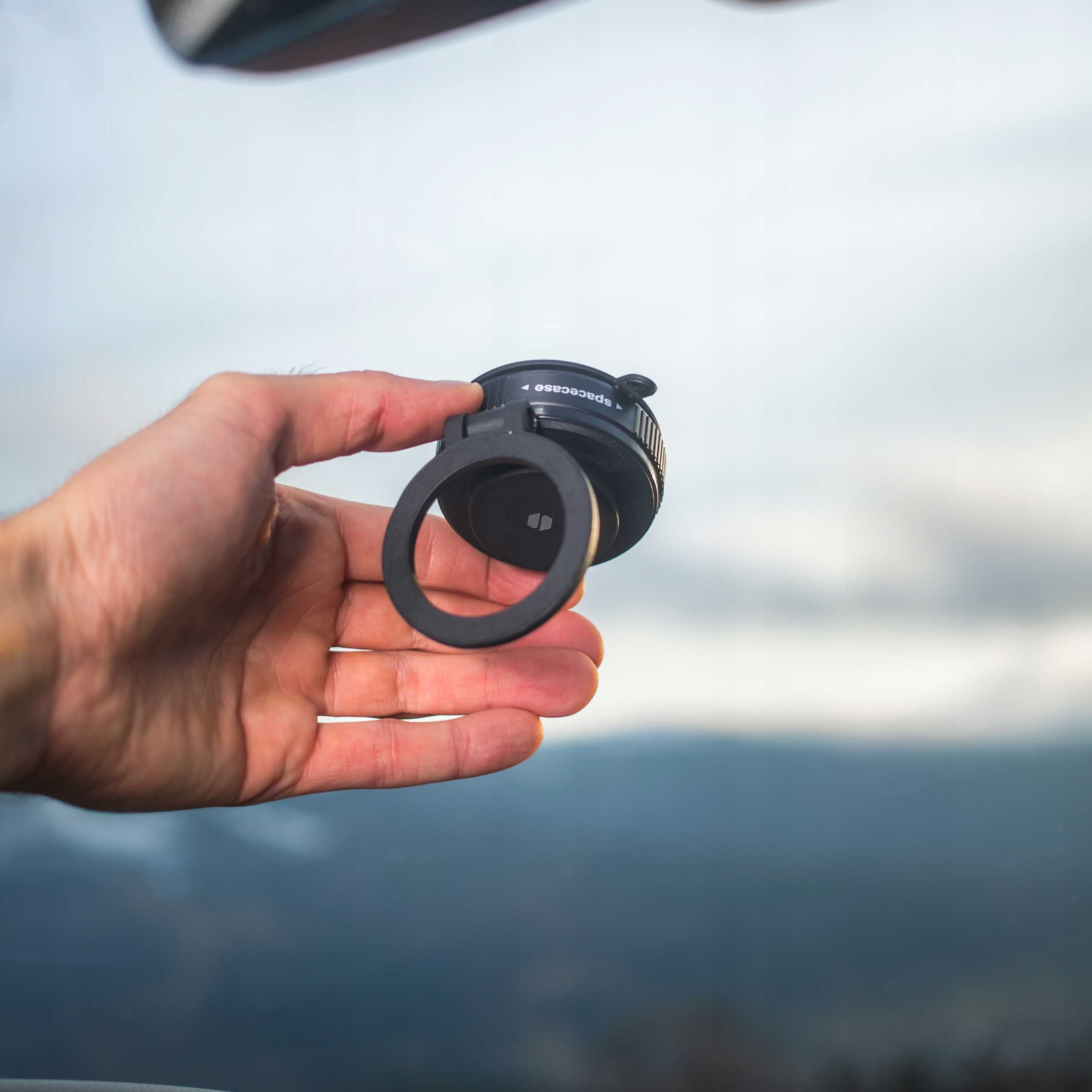 Ręka trzyma czarne urządzenie Spacecase z pierścieniem. Tło pokazuje góry i pochmurne niebo.