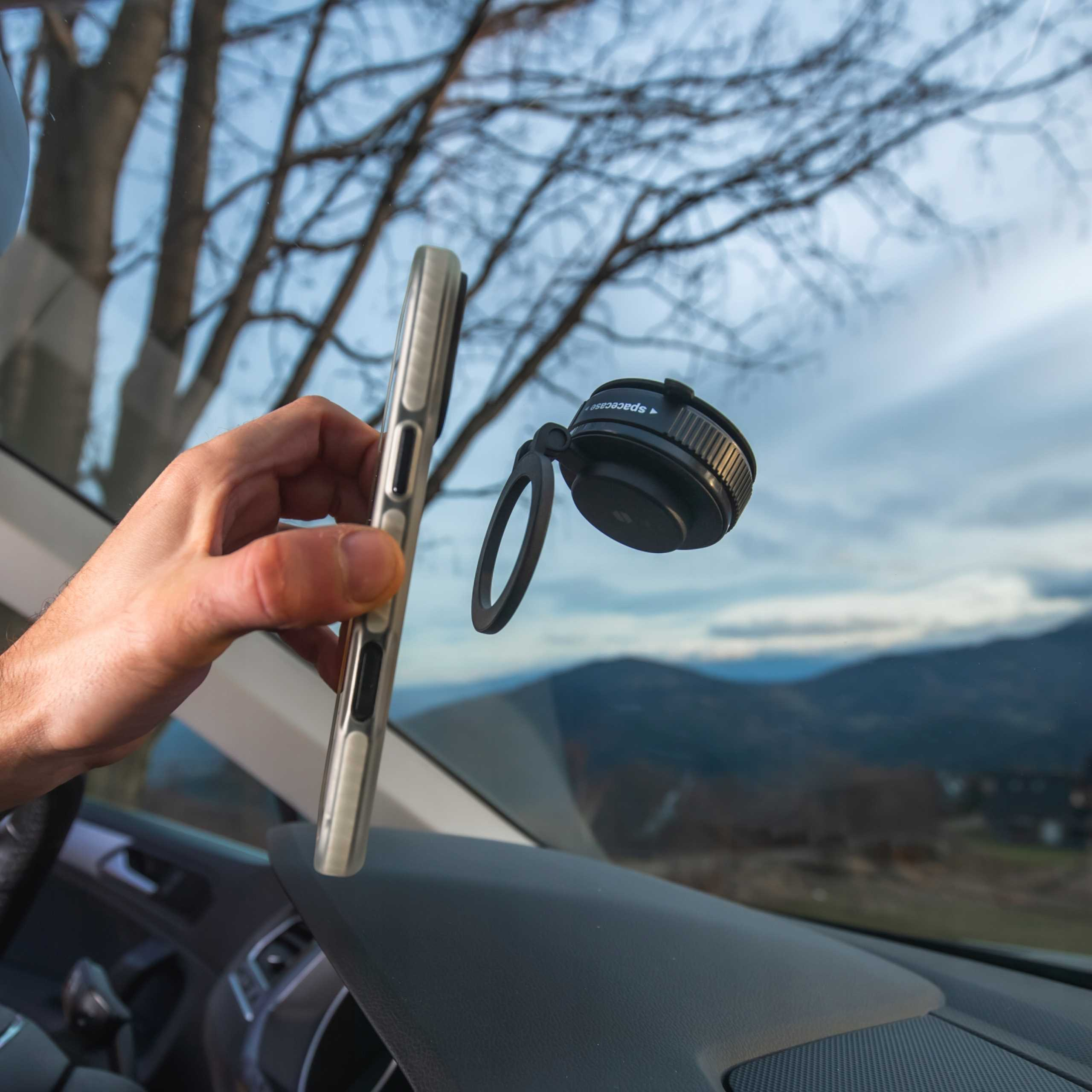 Ręka trzymająca telefon w pobliżu uchwytu samochodowego. Tło: góry i niebo przez przednią szybę.