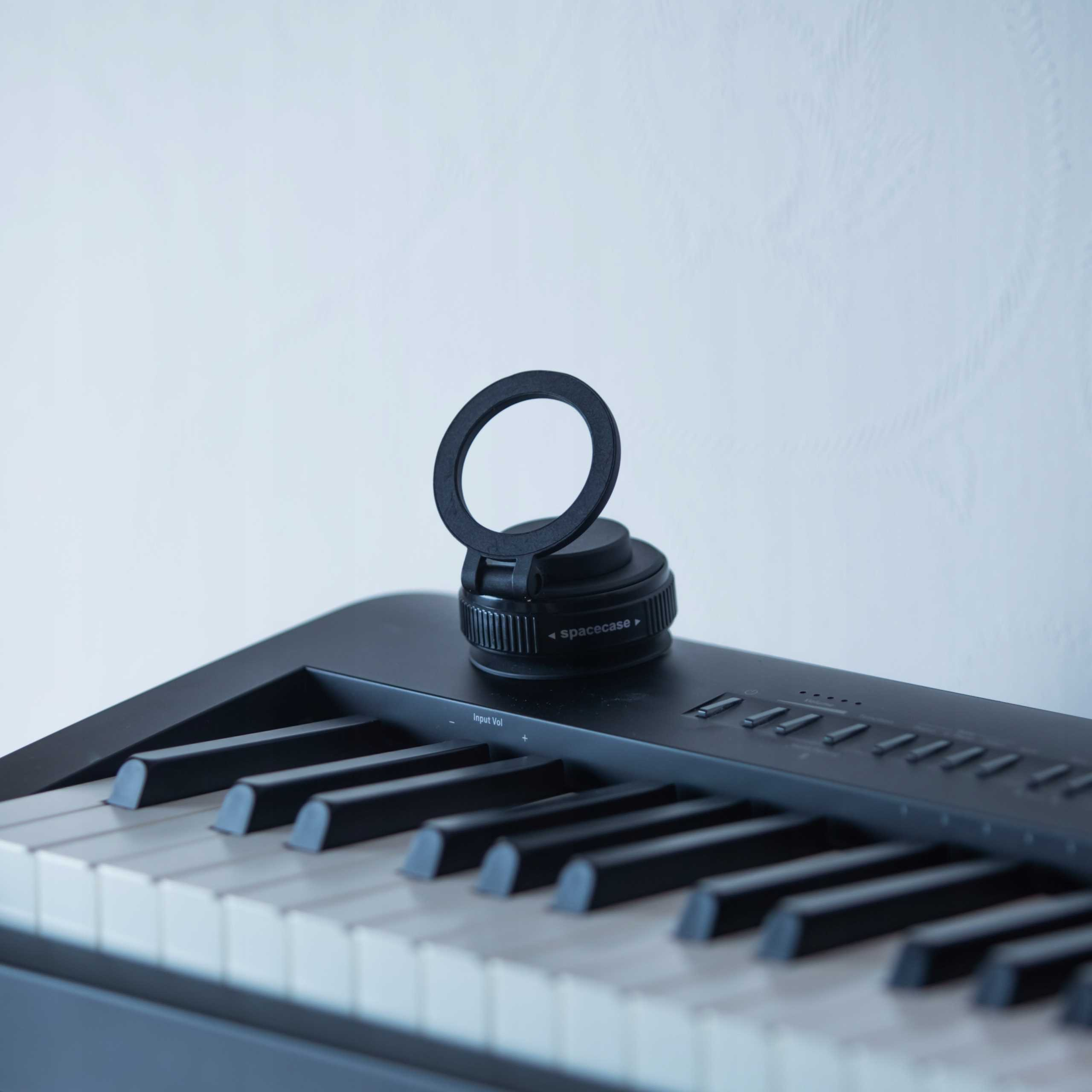 Czarna klawiatura z białymi klawiszami. Na niej czarny okrągły przedmiot. Tło to gładka ściana.