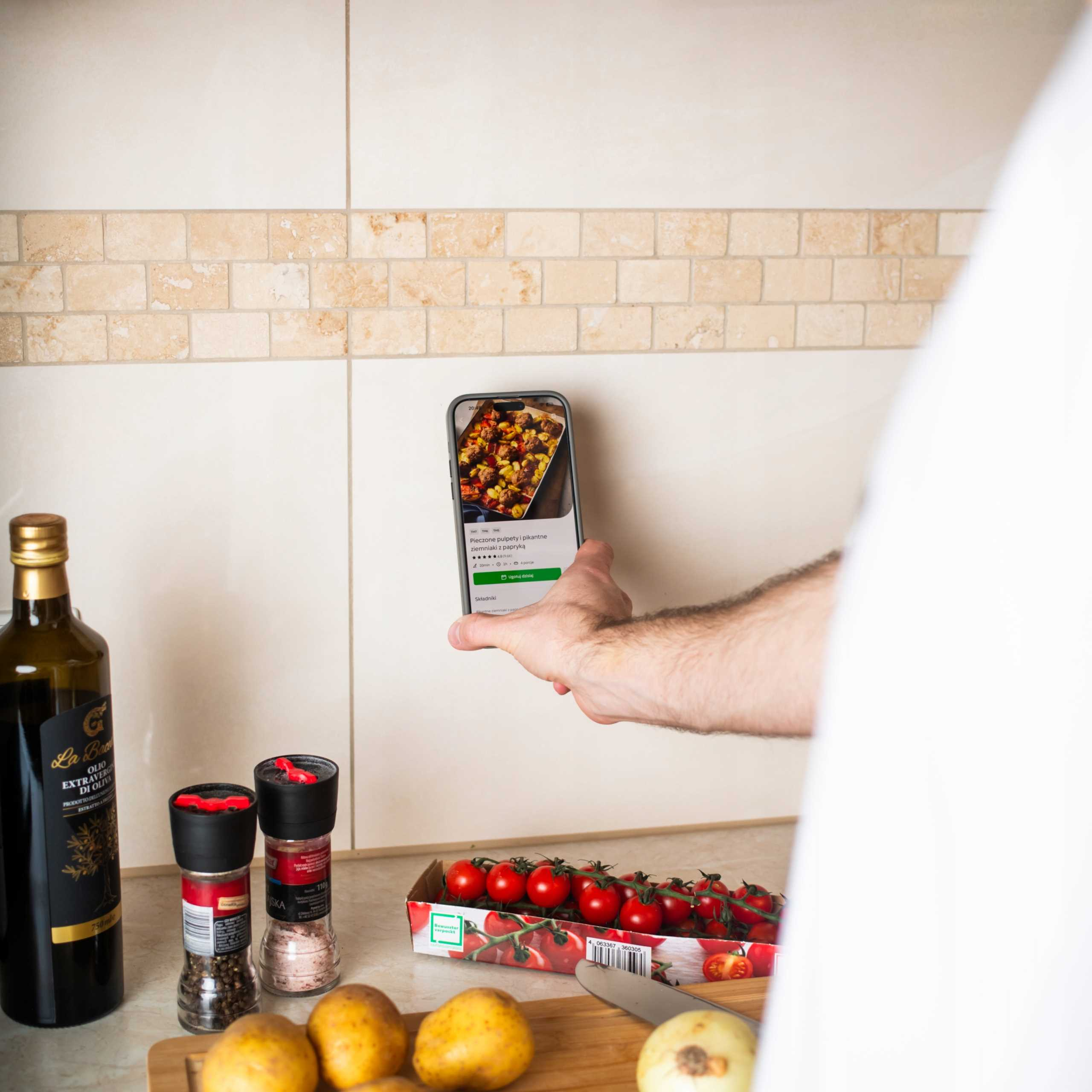 Ręka trzyma telefon z przepisem. Pomidory, ziemniaki, olej i przyprawy na blacie.