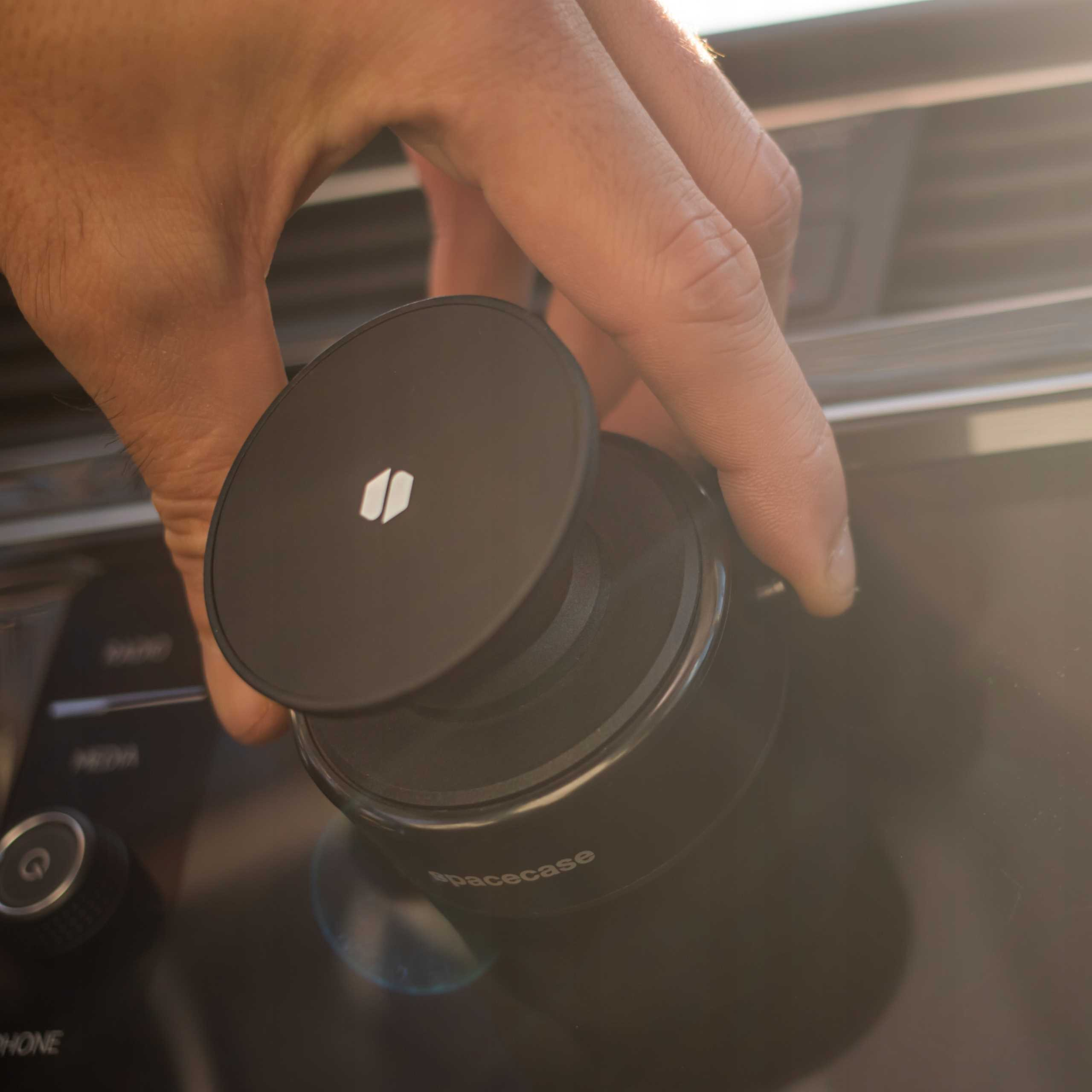 Ręka umieszcza czarne urządzenie Spacecase na konsoli samochodowej. Urządzenie ma białe logo. Tło jest rozmyte.