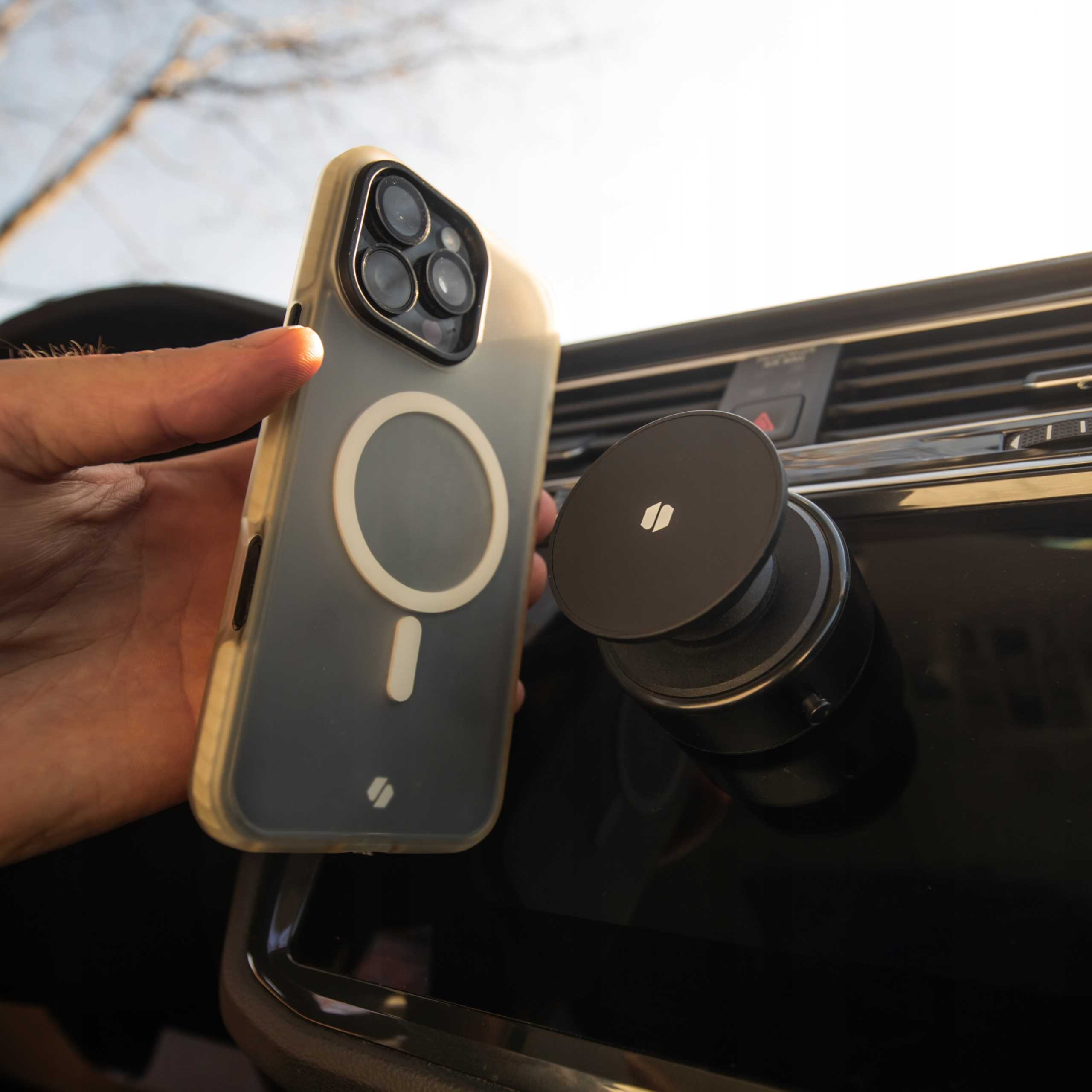 Ręka trzymająca telefon w etui z uchwytem samochodowym. Scena rozgrywa się w samochodzie, ze świecącym słońcem.