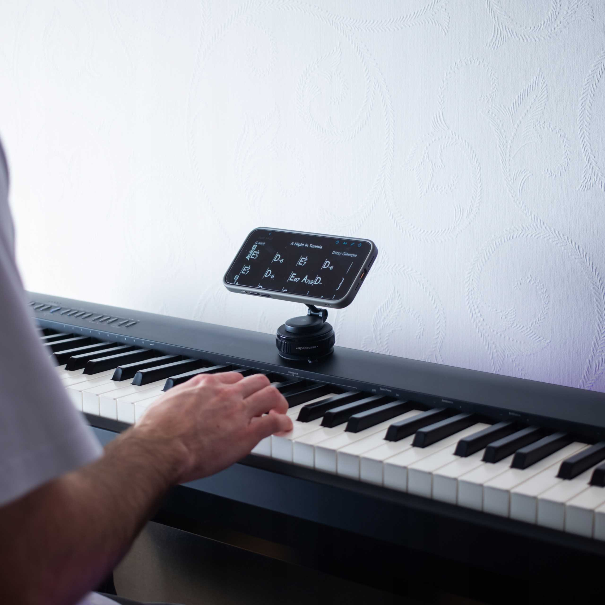 Osoba gra na czarnym pianinie. Telefon wyświetla nuty. Biała, teksturowana ściana w tle.