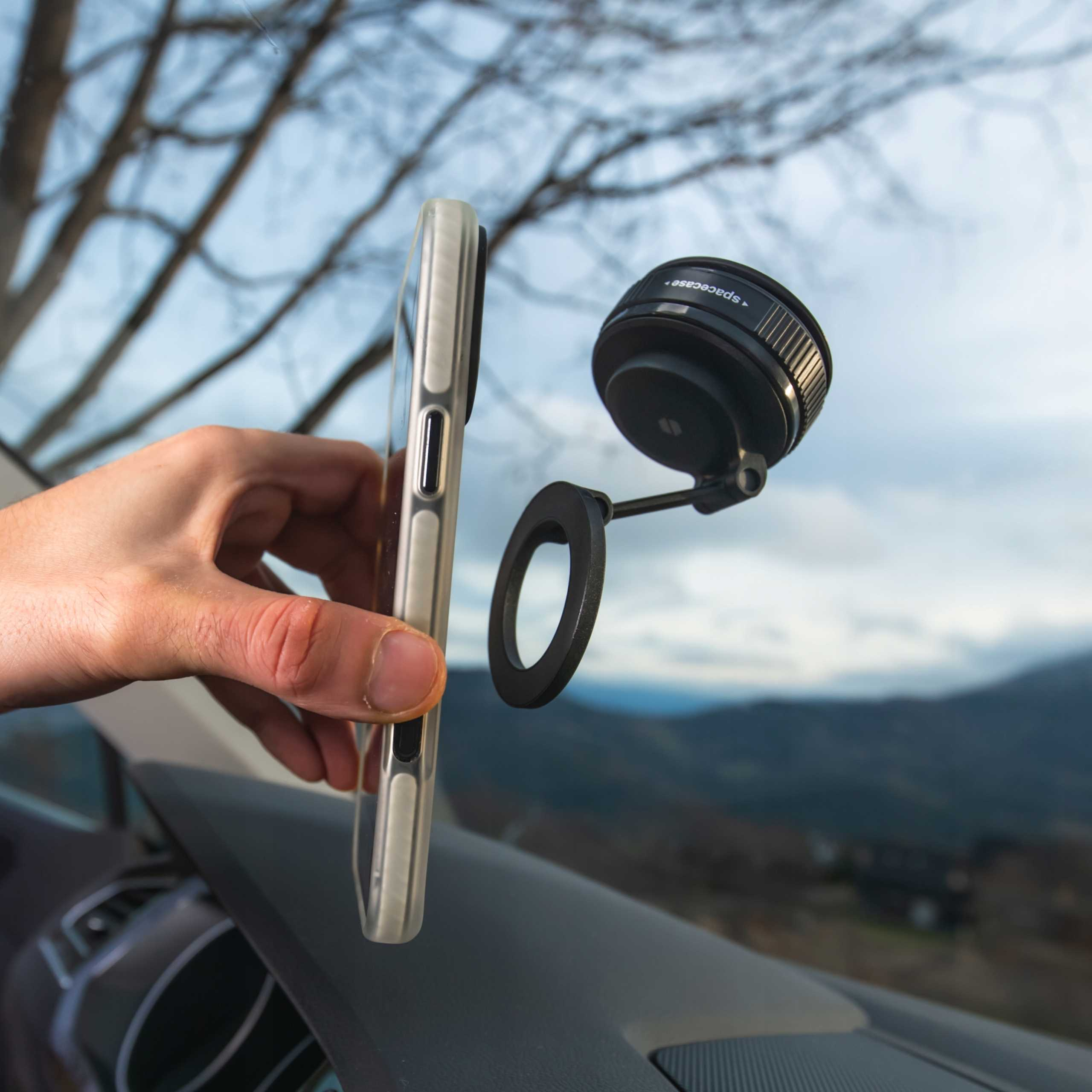 Ręka trzymająca telefon z etui, obok mocowania obiektywu. W tle góry i drzewa, na pierwszym planie deska rozdzielcza samochodu.