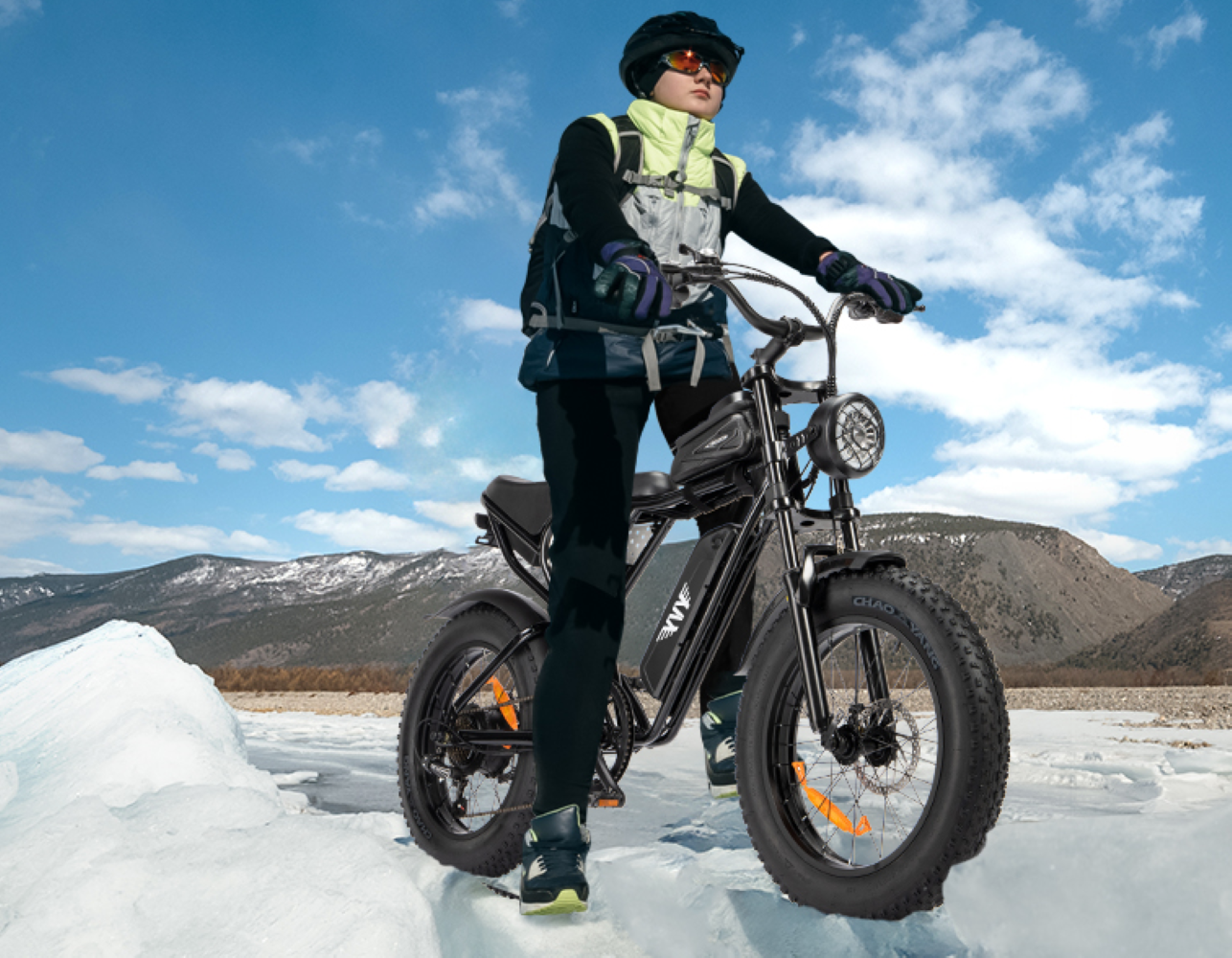 Persona en bicicleta eléctrica negra sobre hielo. Montañas y cielo azul al fondo. La persona lleva casco y equipo de protección.