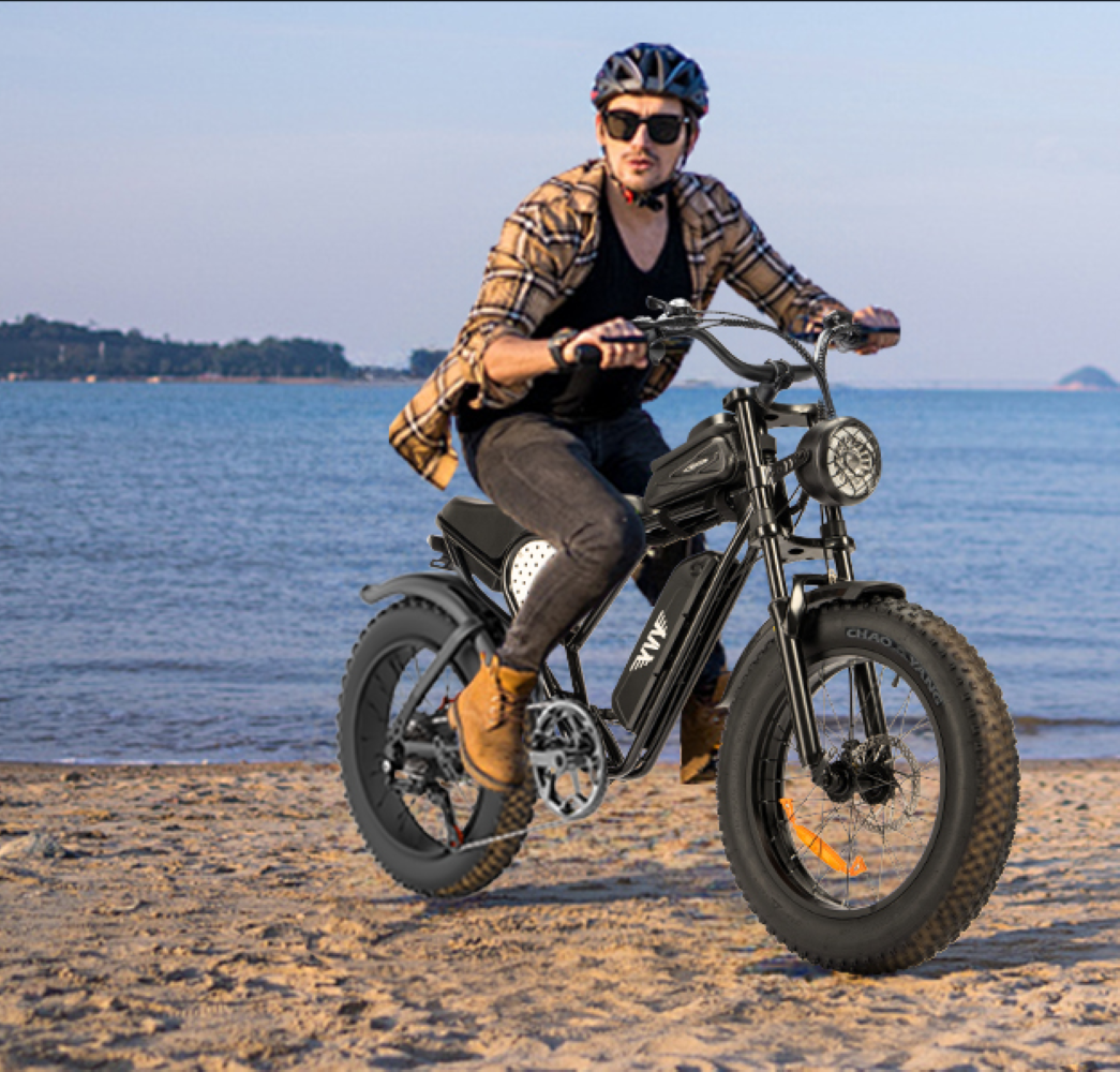 Hombre montando una bicicleta eléctrica negra en la playa. Lleva casco y gafas de sol. El mar está al fondo.
