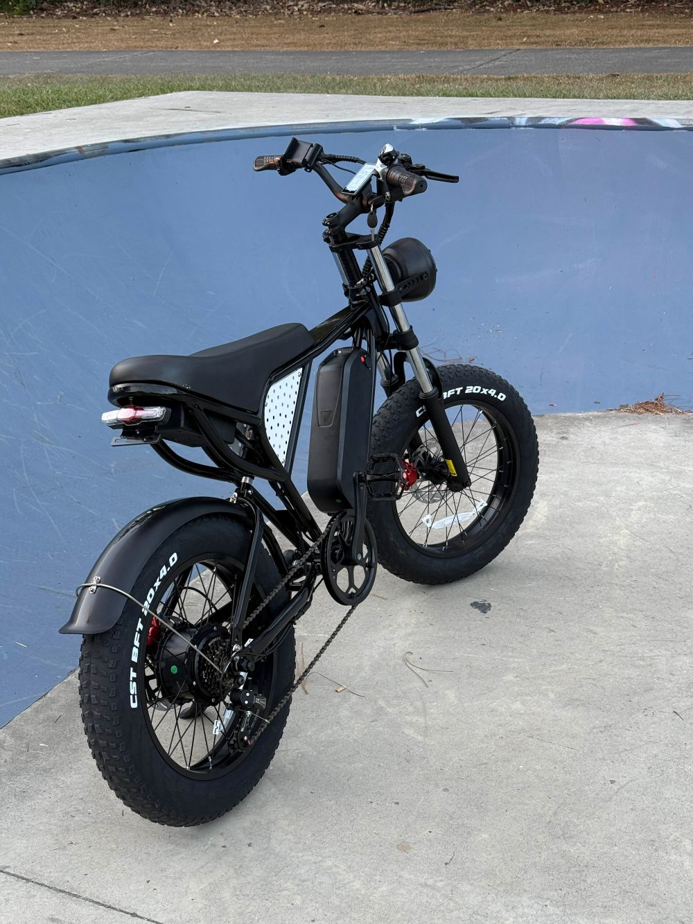 Bicicleta eléctrica negra sobre hormigón. Skate park azul al fondo. La bicicleta tiene neumáticos grandes y batería.