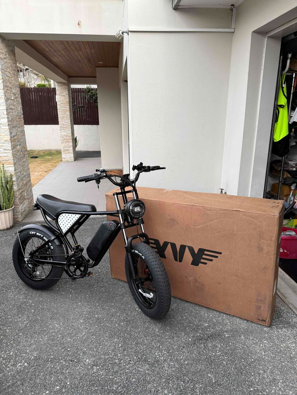 Bicicleta eléctrica negra estacionada junto a una gran caja de cartón. La bicicleta tiene neumáticos gruesos, un cuadro negro y un asiento blanco con puntos.