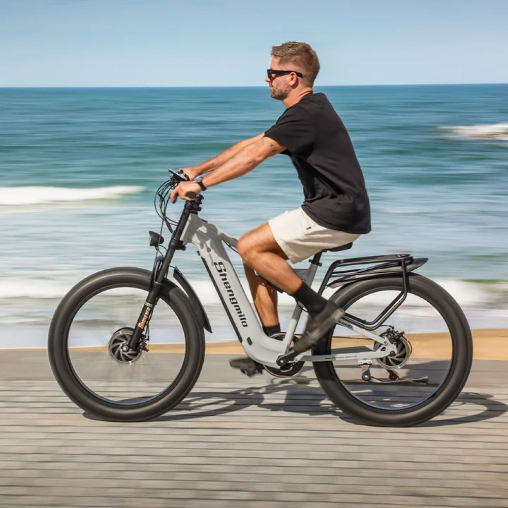 Un hombre con gafas de sol monta una bicicleta eléctrica gris sobre un camino de madera. El mar está al fondo en un día soleado.