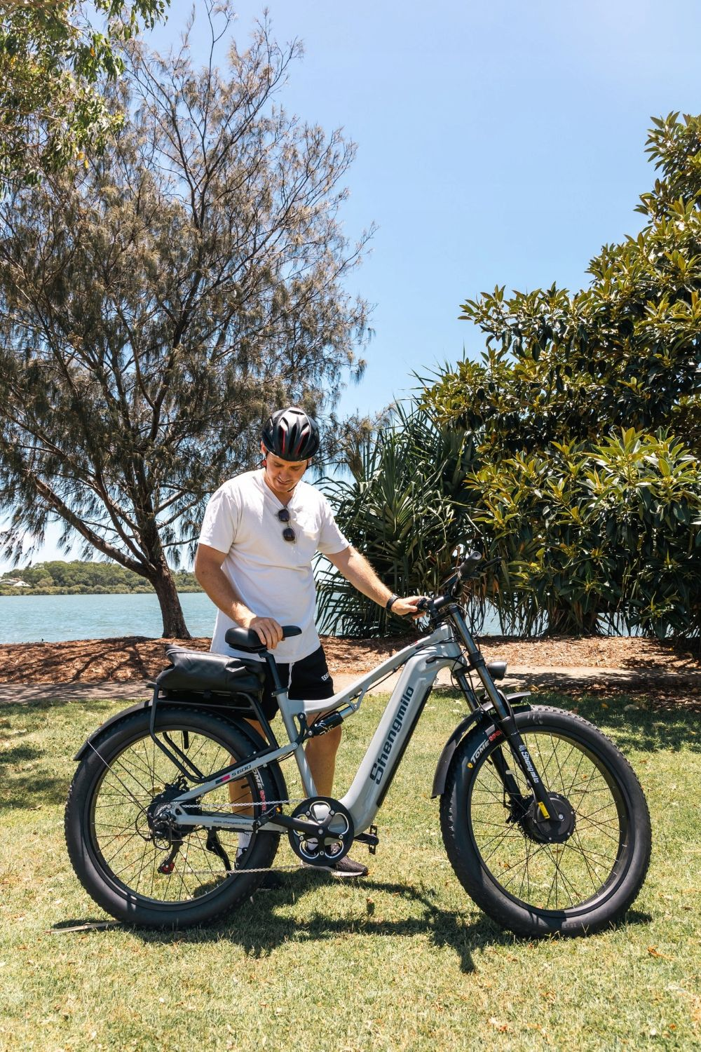 Un hombre con casco está junto a una e-bike gris Shengmilo sobre césped. Árboles y un lago al fondo bajo cielo azul.