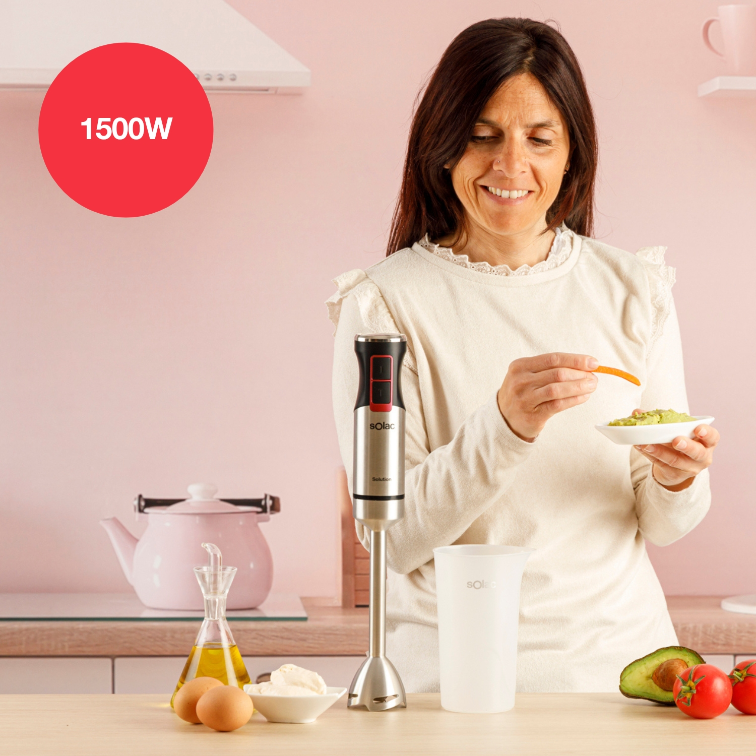 Una mujer prepara comida con una batidora, sosteniendo una zanahoria sobre un plato de guacamole. Encimera de cocina con ingredientes.