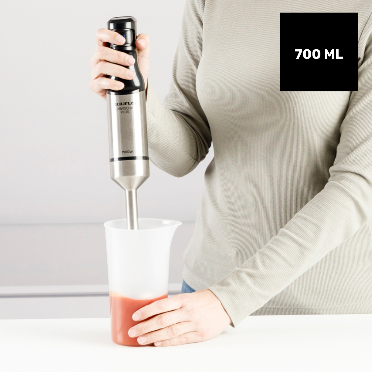 Una persona usa una batidora de mano para mezclar un líquido rojo en un recipiente blanco. La batidora es plateada y negra, con etiqueta 700ml.