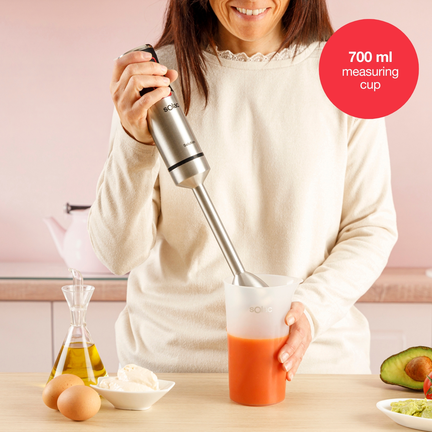 Mujer usa una batidora en una taza medidora con líquido naranja. Huevos, aceite y aguacate en la mesa.