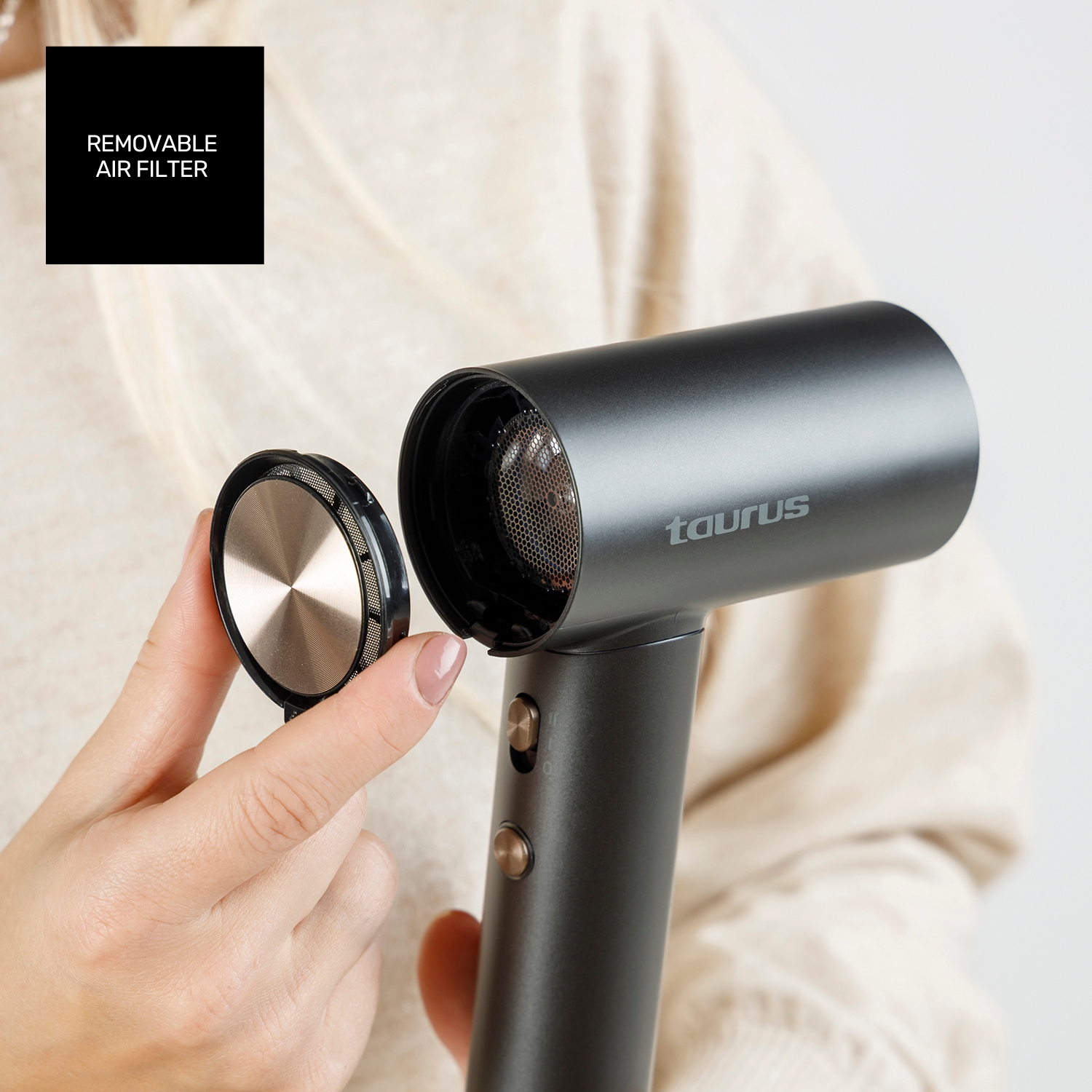 Una persona sostiene un secador de pelo con un filtro de aire extraíble. El secador es gris con detalles dorados. Se está quitando el filtro.