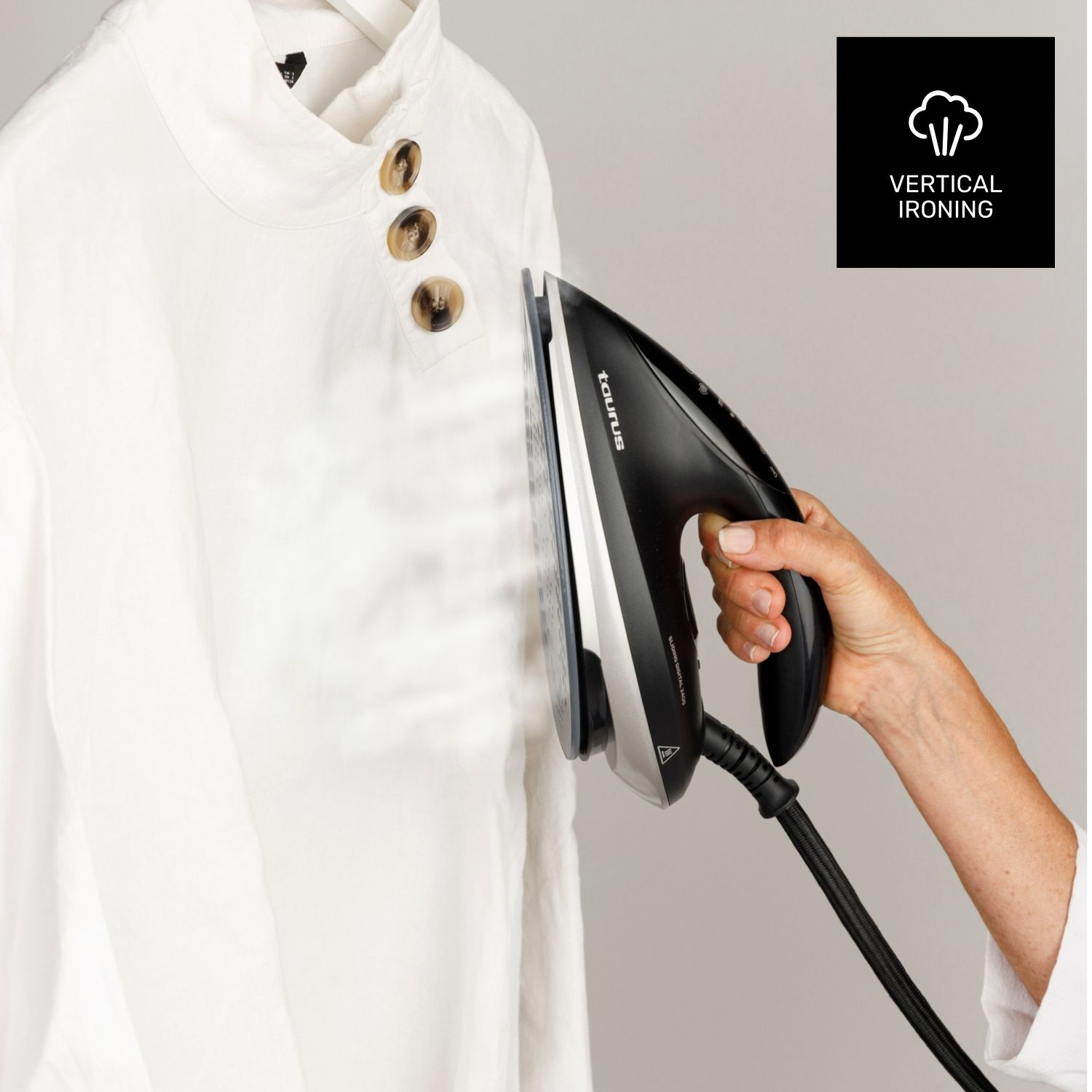 Una mano stira una camicia bianca con un ferro da stiro nero e argento. Il vapore sale. I bottoni sono visibili sulla camicia. Lo sfondo è grigio.