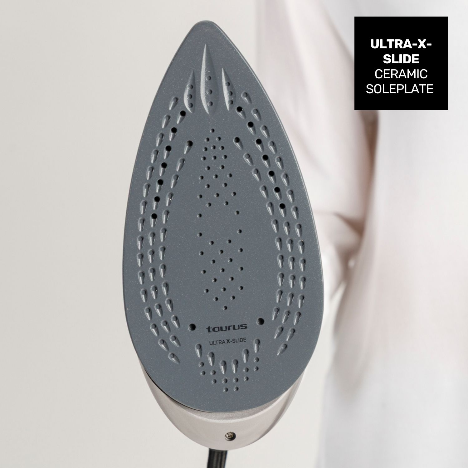 Primo piano di una piastra di ferro grigia con fori. Il ferro è tenuto da una persona. Testo: ULTRA-X-SLIDE CERAMIC SOLEPLATE.