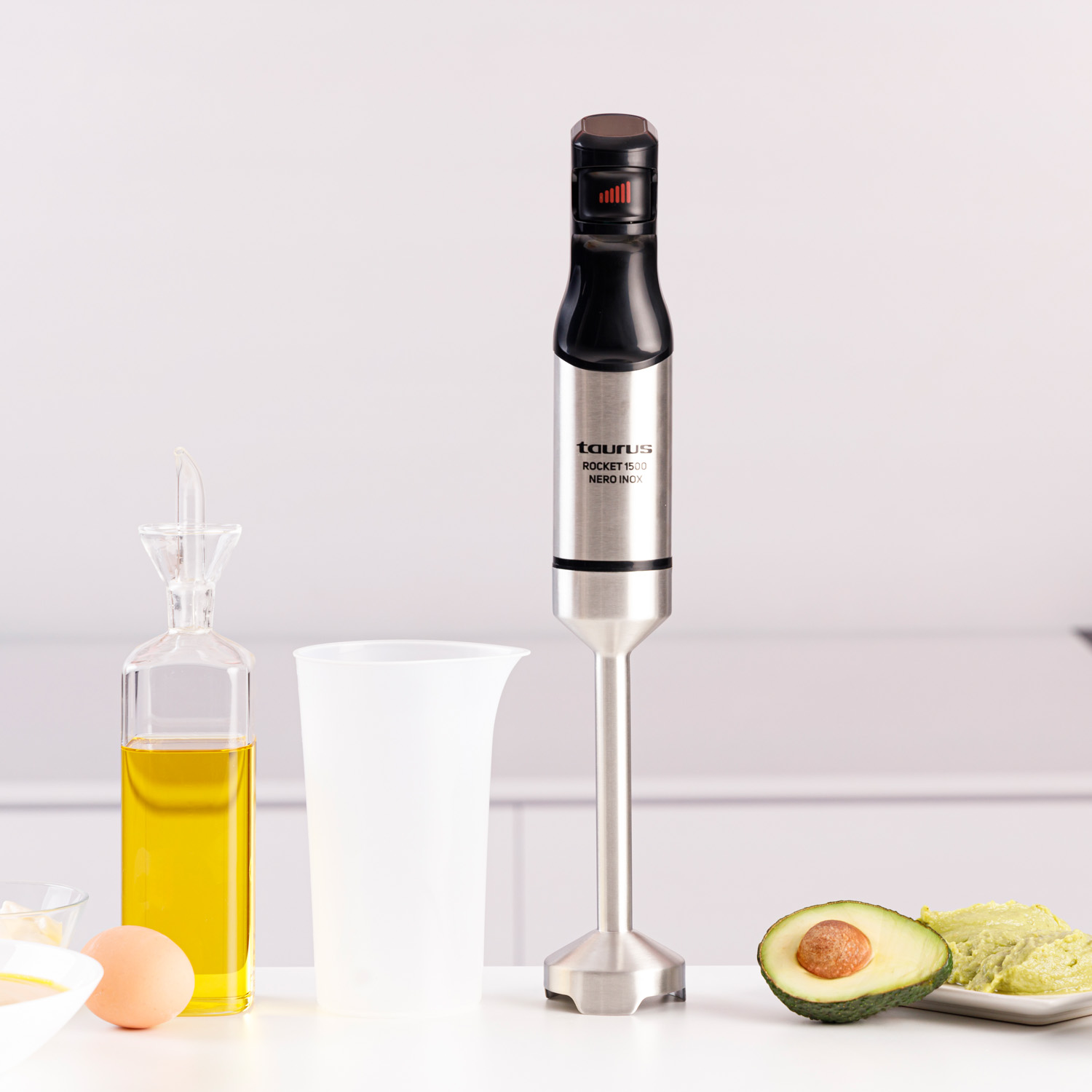 Una batidora de mano con ingredientes: aceite, huevo, aguacate y una taza medidora. La batidora está en el centro.