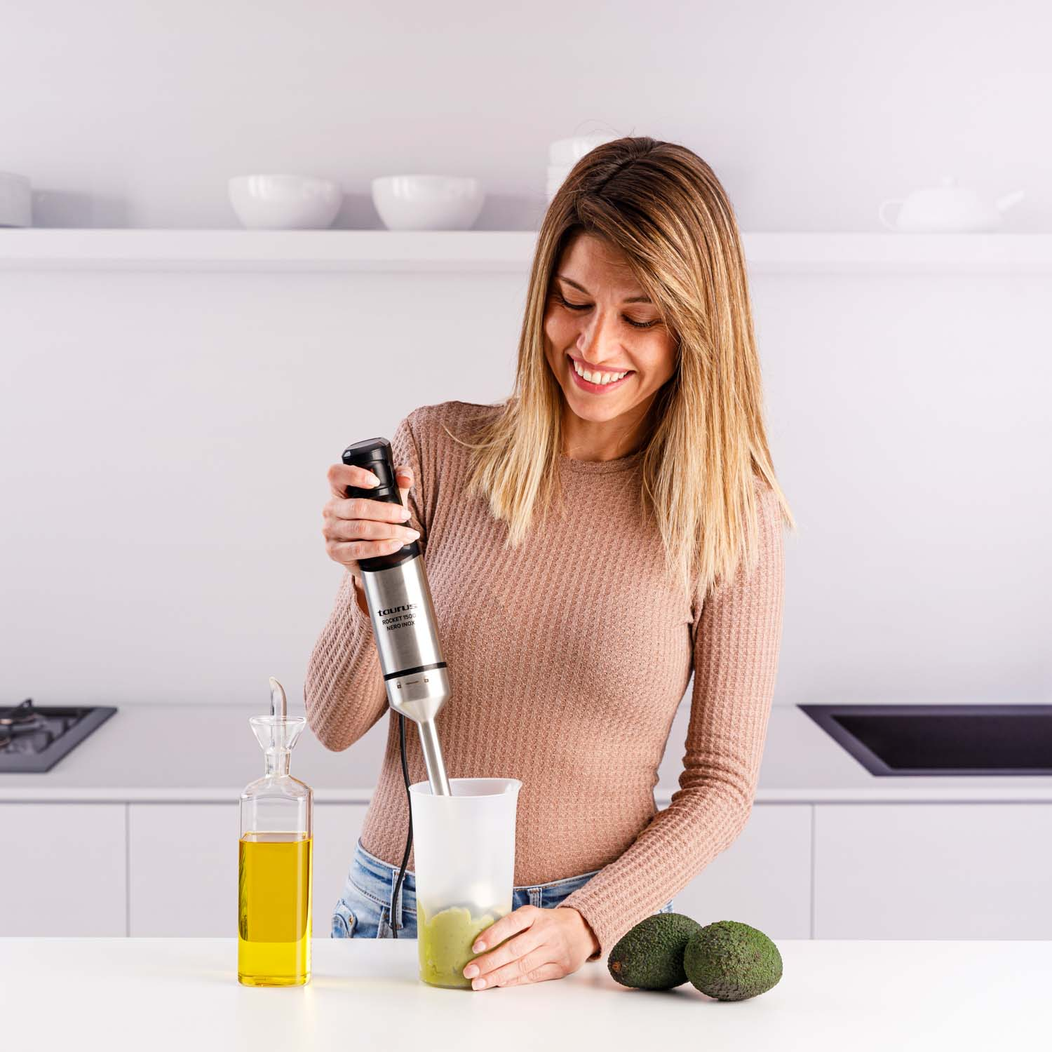 Mujer sonríe, usando una batidora de mano. Mezcla de aguacate en una taza, aguacates y botella de aceite en la mesa blanca. Fondo de cocina.