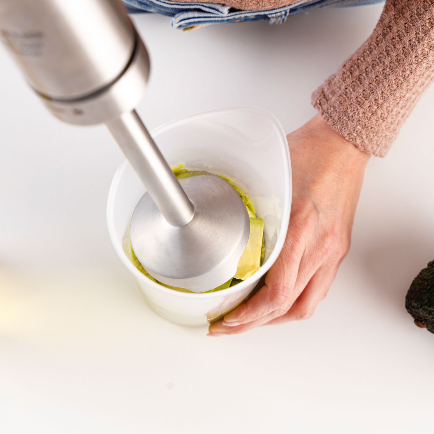 Una persona usa una batidora de inmersión para mezclar aguacate en un recipiente blanco. La escena está en una superficie blanca con un aguacate.