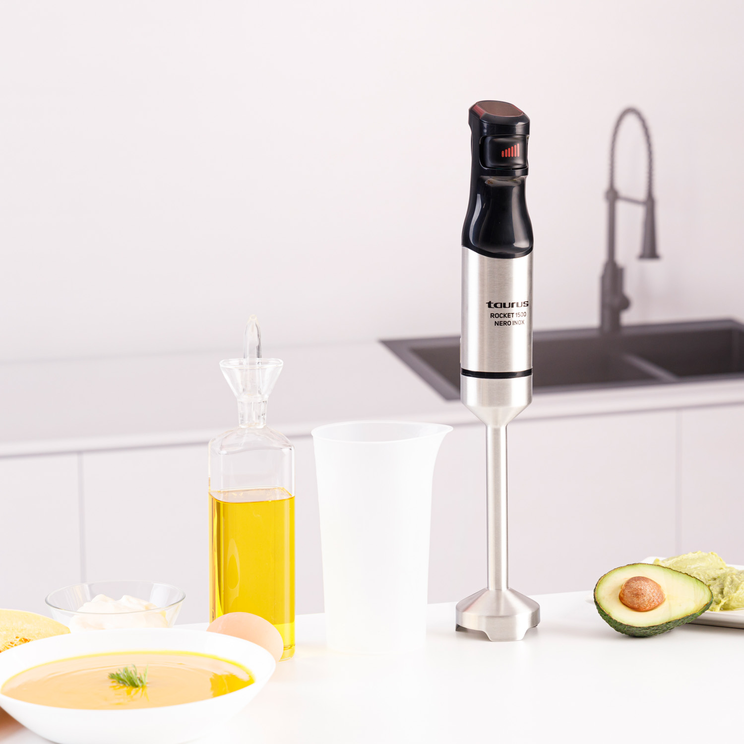 Una escena de cocina con una batidora, aceite, sopa, huevo, aguacate y una taza medidora sobre una superficie blanca. El fondo muestra un fregadero.