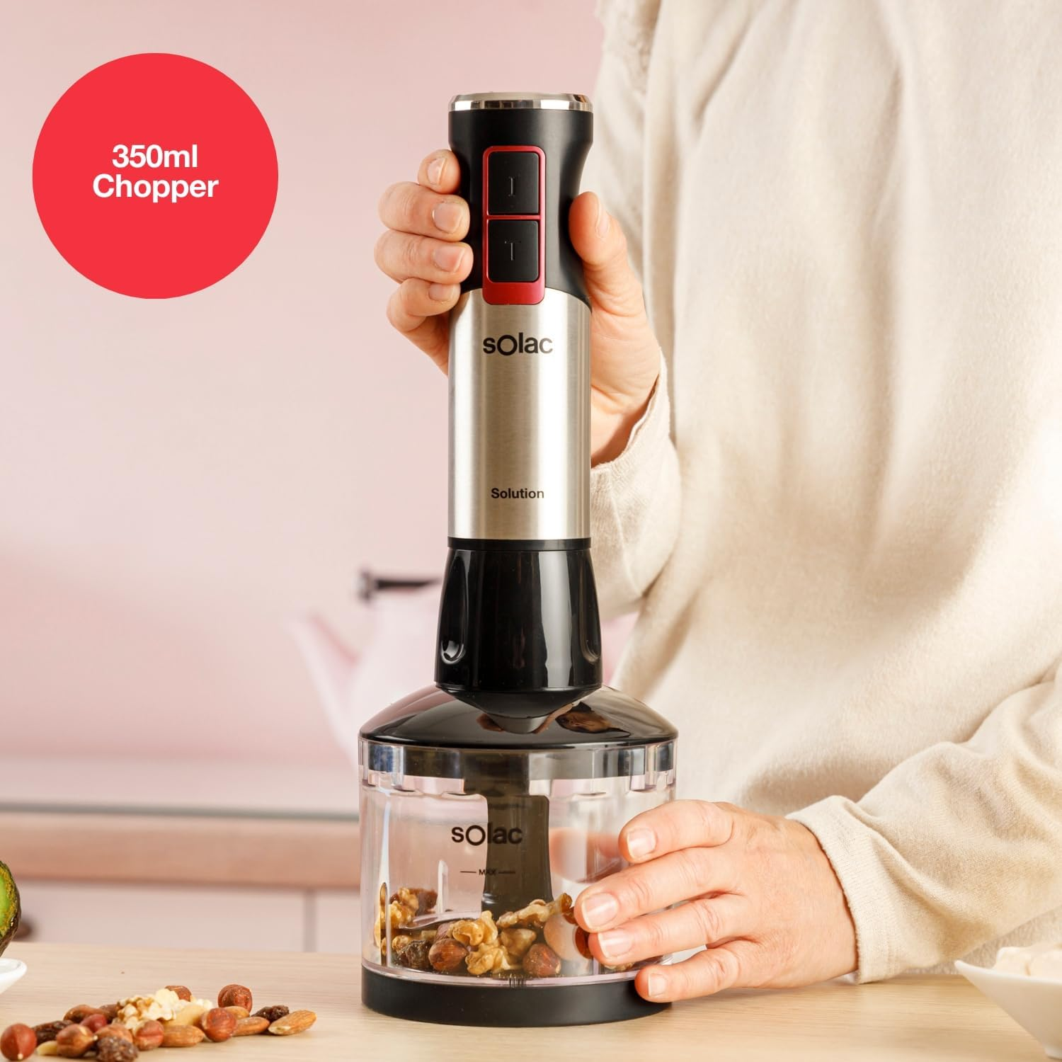 Una persona usa una batidora de mano Solac. La batidora es plateada y negra, con un recipiente transparente lleno de nueces. Un círculo rojo dice 350ml Chopper.
