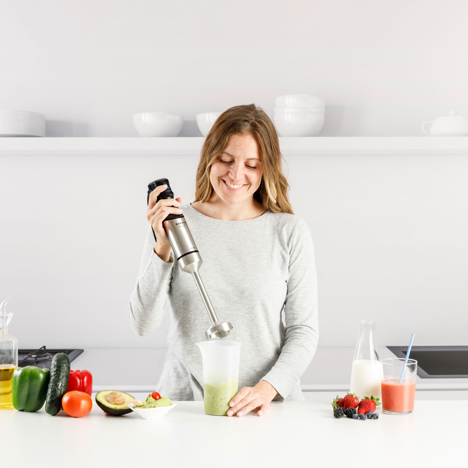 Mujer sonríe mientras mezcla un batido. Verduras, frutas y jugo en la mesa. Fondo de cocina blanco.