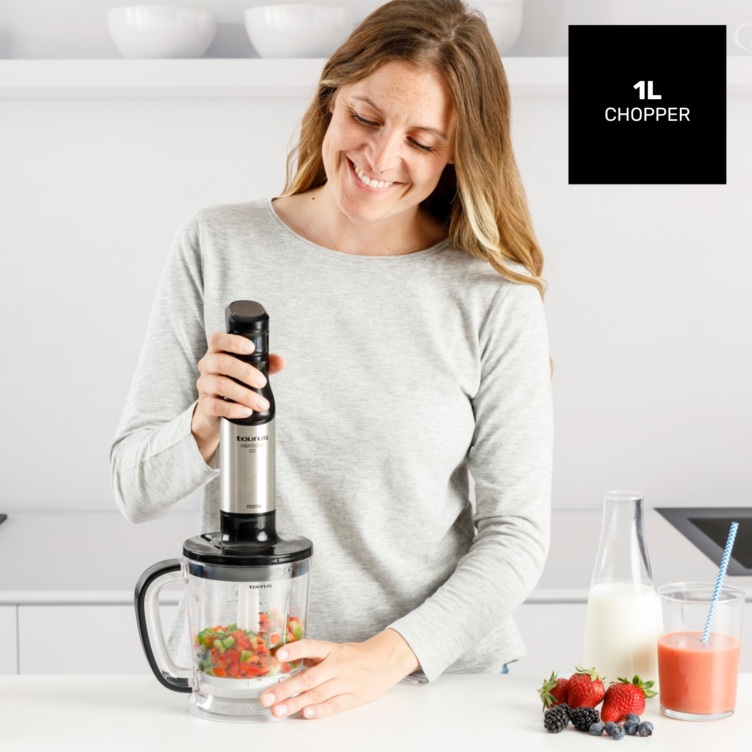 Una mujer usa una licuadora para picar verduras. Frutas, batidos, leche y bayas están en la mesa. Un picador de 1L está en el fondo.