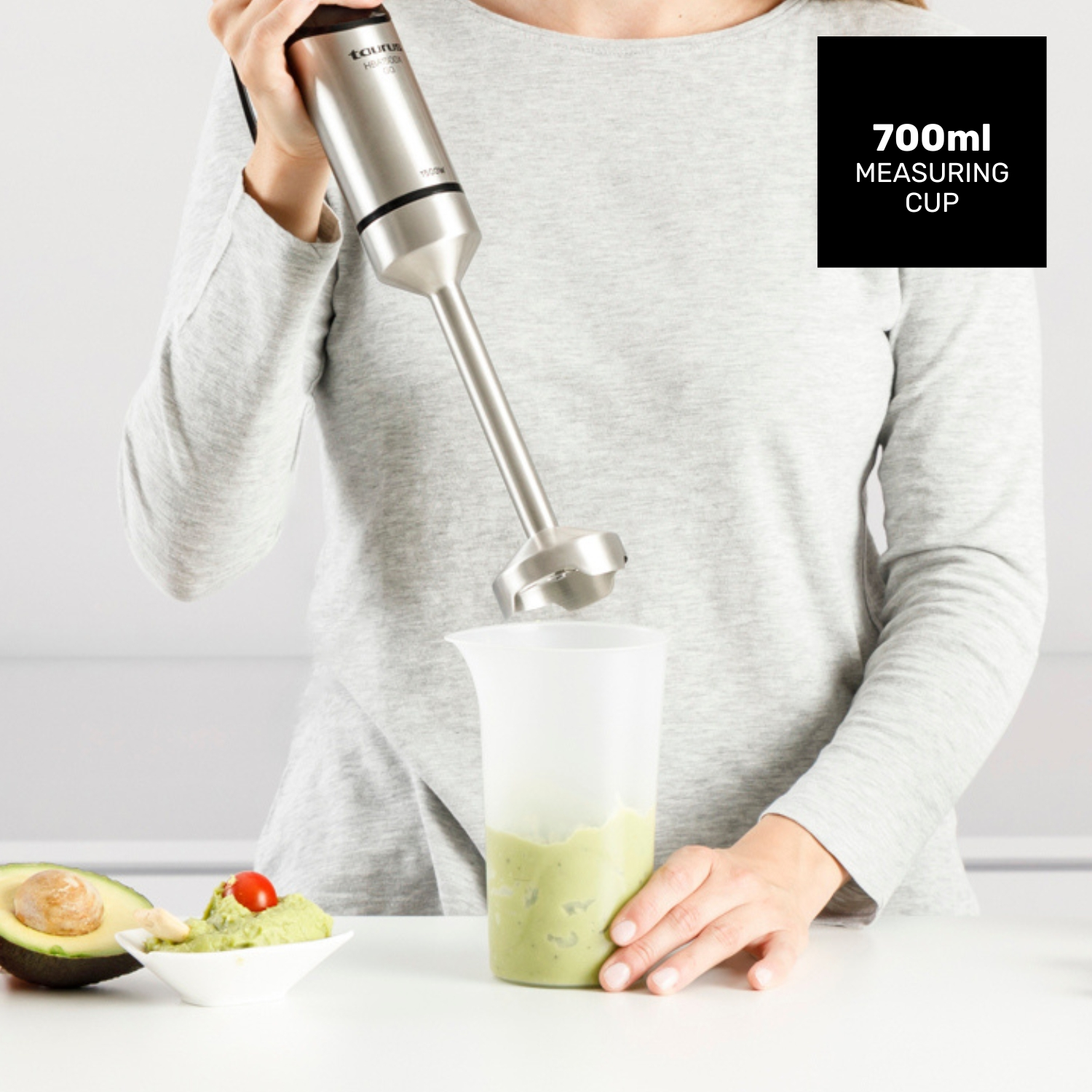 Una mujer mezcla aguacate en una taza medidora con una batidora de mano. Mitad de aguacate y guacamole en una mesa.