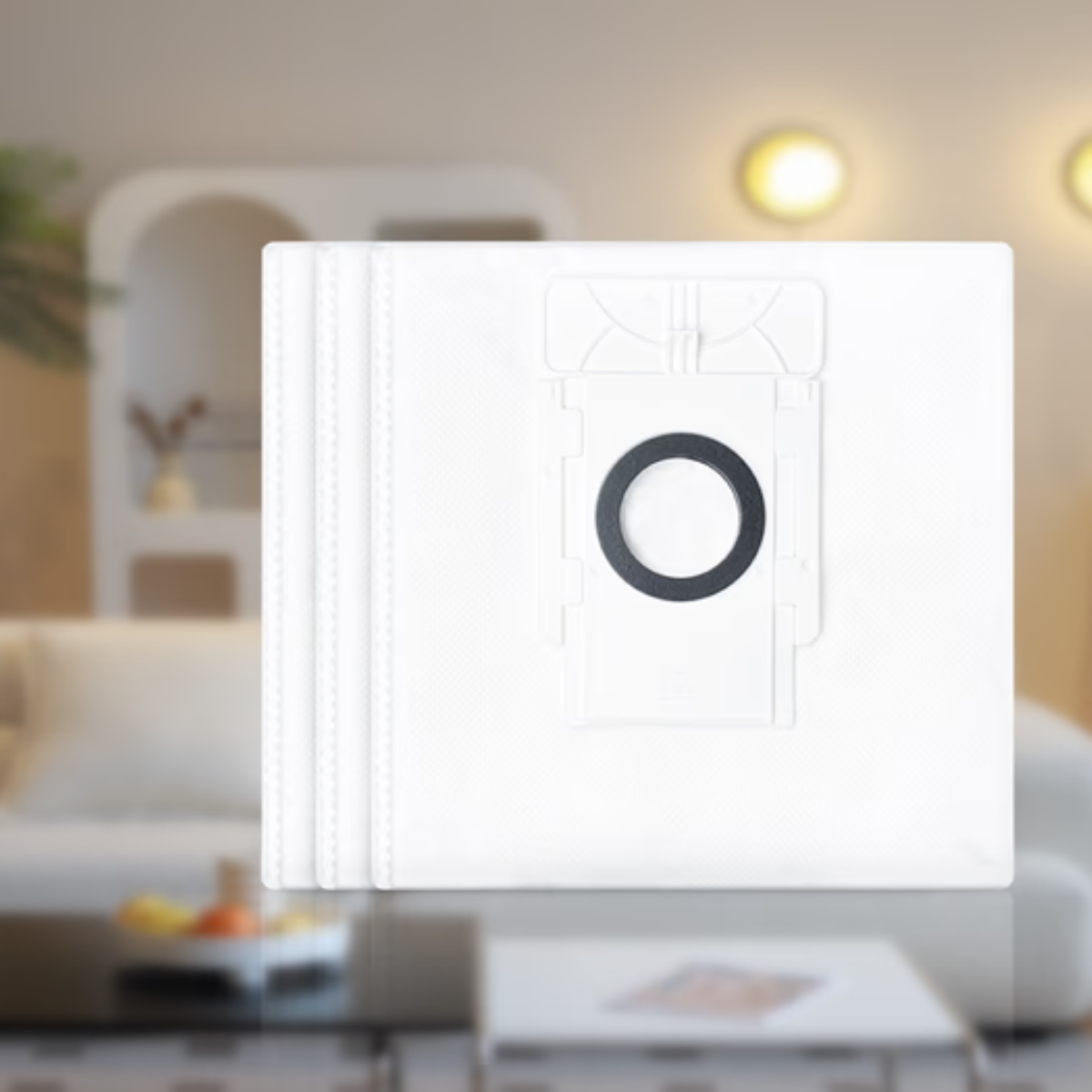 Biały worek do odkurzacza z czarnym pierścieniem, ułożony w stos. Rozmyte tło przedstawia salon z sofą i stolikiem kawowym.