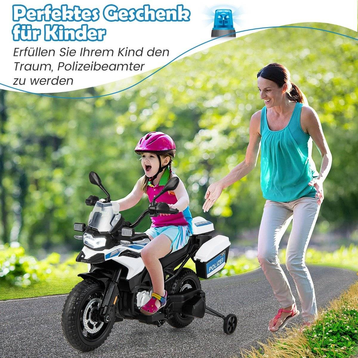 Una niña en una moto de policía de juguete es conducida por una mujer en una carretera. Están en un parque con hierba verde y árboles de fondo.