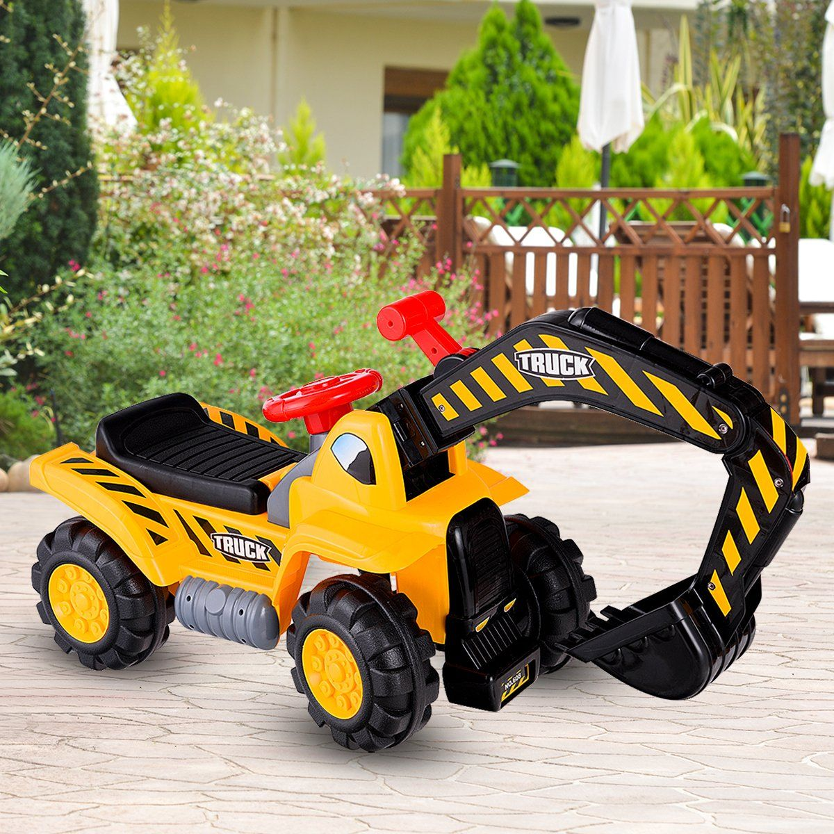 Un camión de juguete amarillo con rayas negras y amarillas. Tiene un volante rojo y un brazo excavador negro. El fondo es un jardín.