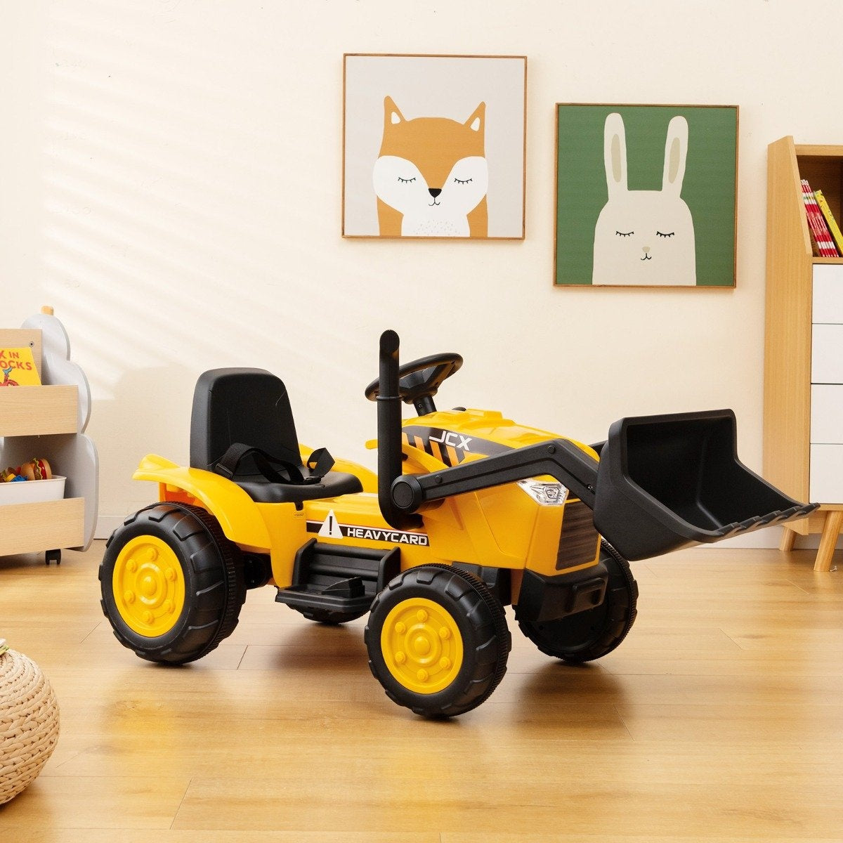 Un tractor de juguete amarillo con neumáticos negros y una pala delantera está sobre un piso de madera. Dos retratos de animales enmarcados cuelgan en la pared.