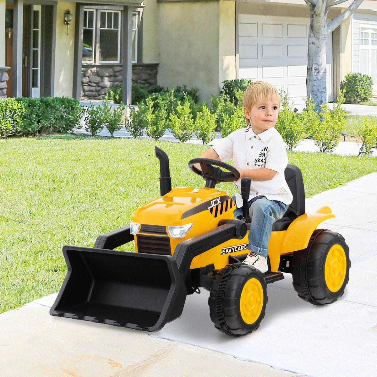 Un niño pequeño está sentado en un tractor de juguete amarillo con una pala delantera negra. Está afuera frente a una casa en un día soleado.