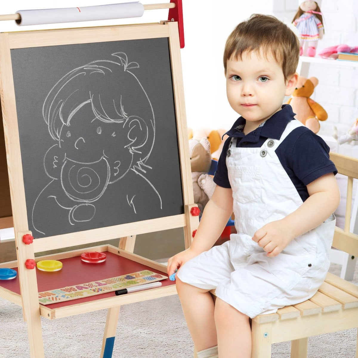 Un niño pequeño se sienta junto a un caballete con un dibujo. Lleva un atuendo blanco y mira a la cámara.