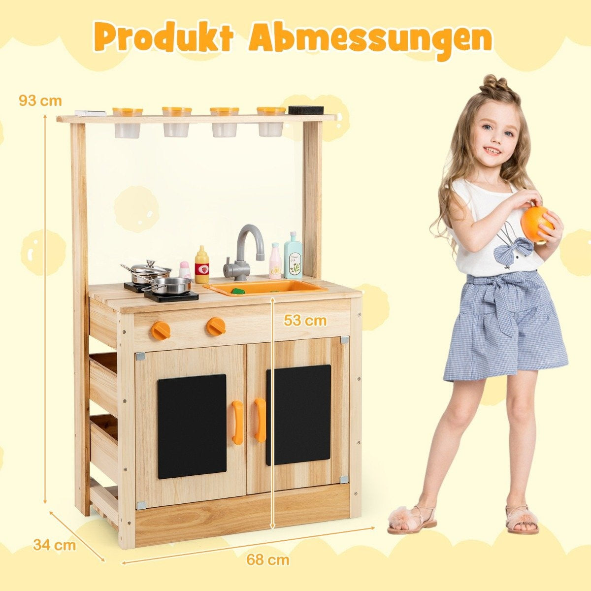 Una niña está junto a una cocina de juguete de madera. La cocina tiene un fregadero, una estufa y armarios. La niña sostiene una naranja.