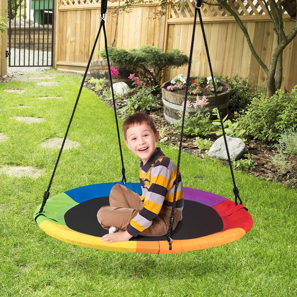 Un niño está sentado en un columpio colorido en un jardín. El columpio es redondo y multicolor. El niño sonríe.