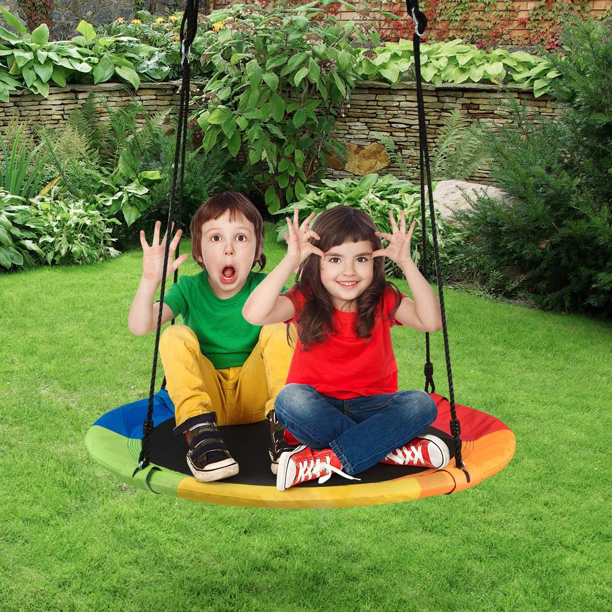 Dos niños sentados en un columpio arcoíris, sonriendo y haciendo gestos con las manos. Césped verde y follaje en el fondo.