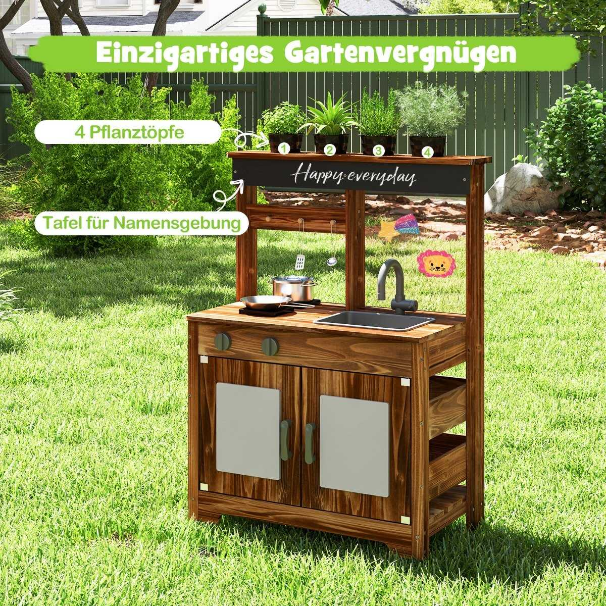 Cocina de juego de madera para exteriores con plantas encima, fregadero y horno. Sobre hierba verde con valla al fondo.