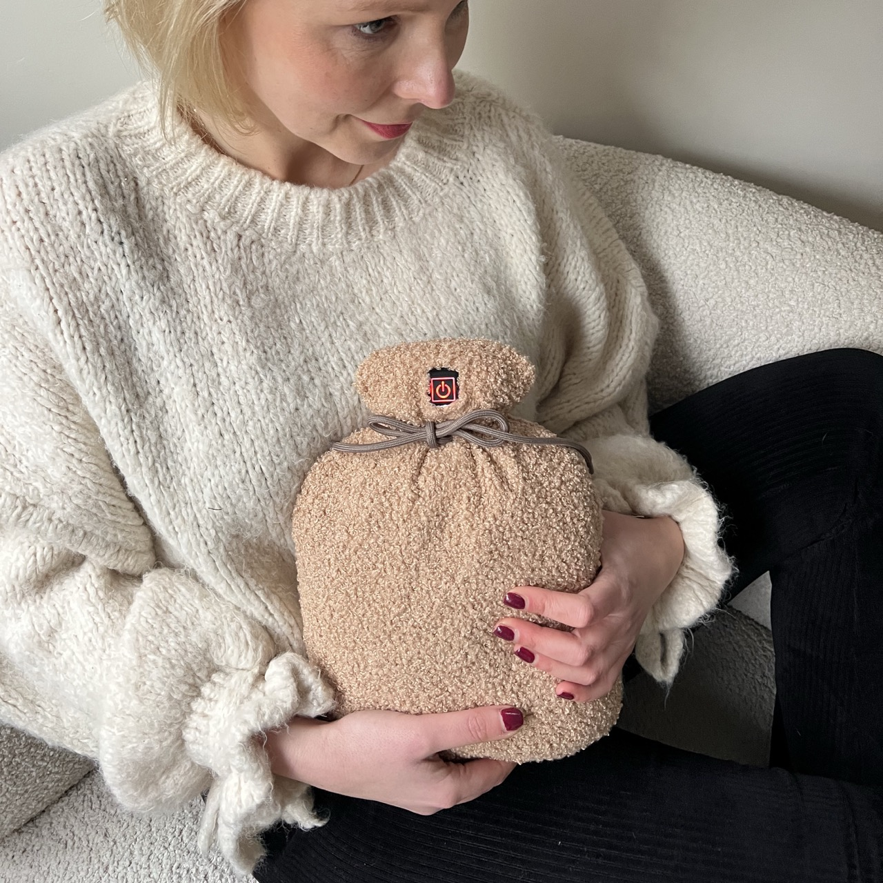 Eine Frau hält eine beige Wärmflasche. Sie trägt einen weißen Pullover und eine schwarze Hose. Ihre Nägel sind rot lackiert.