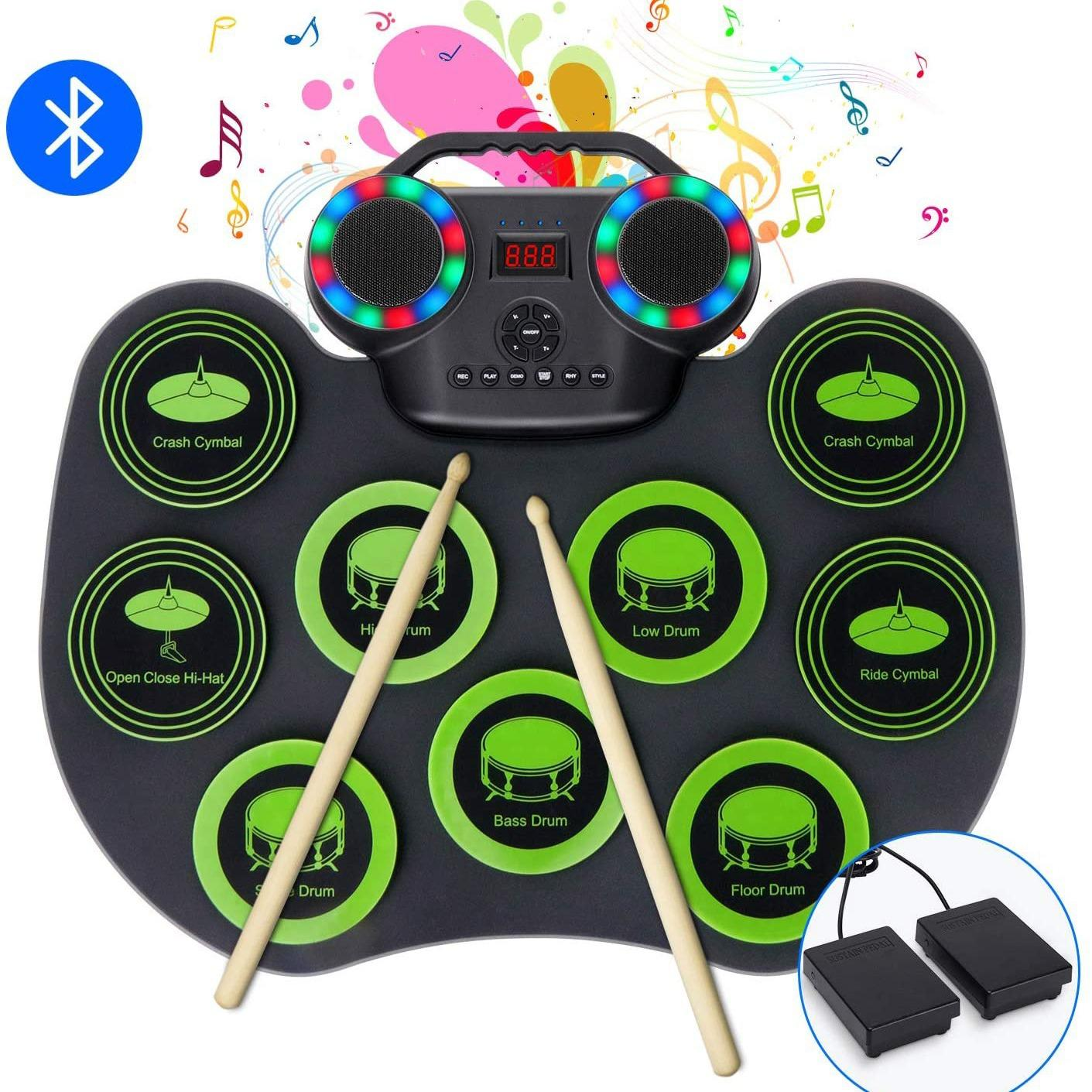 Elektronisches Schlagzeug mit beschrifteten Pads, Trommelstöcken und Fußpedalen. Verfügt über Lautsprecher, Bluetooth und Noten im Hintergrund.