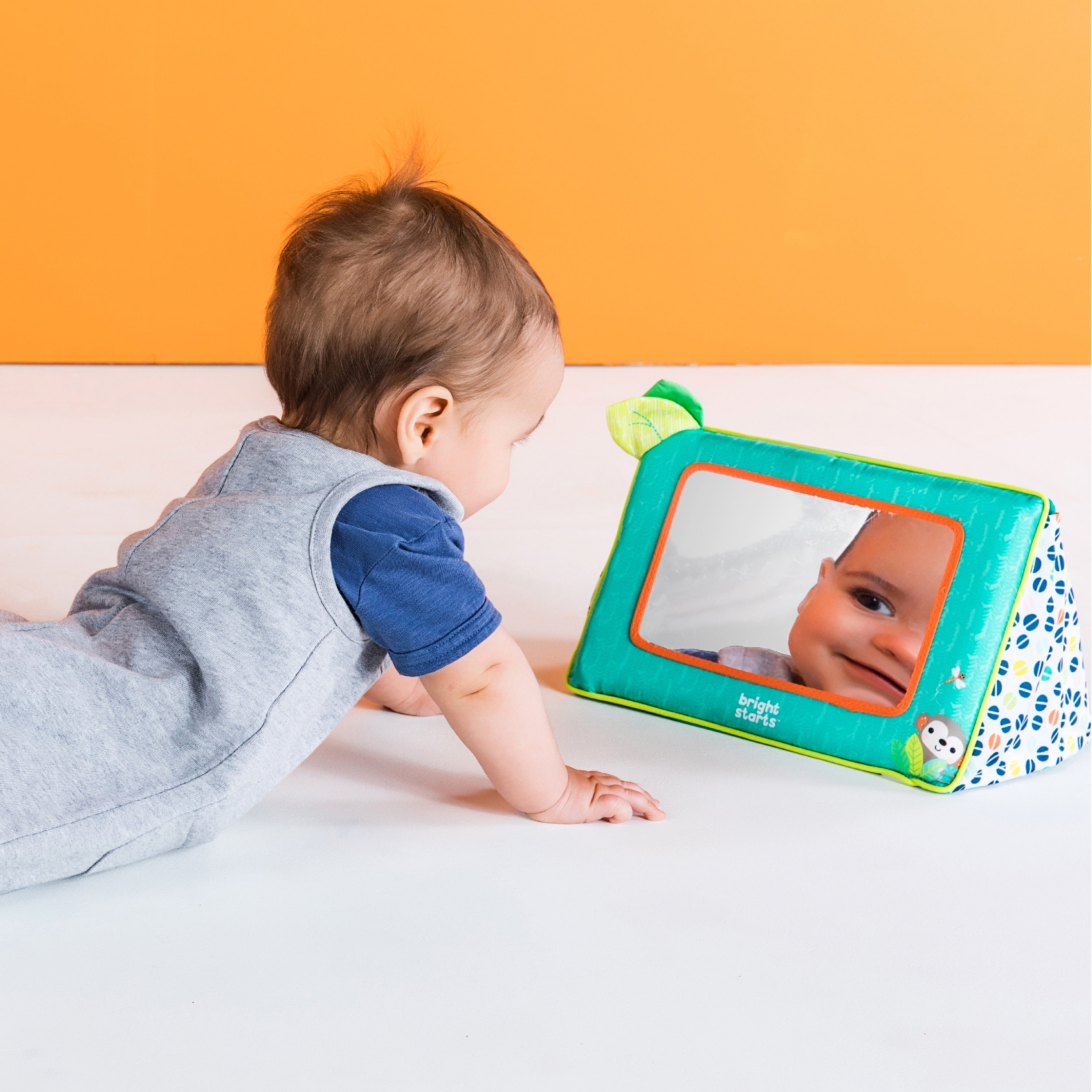 Ein Baby betrachtet ein Spiegelspielzeug. Das Baby trägt graue und blaue Kleidung. Oranger Hintergrund.