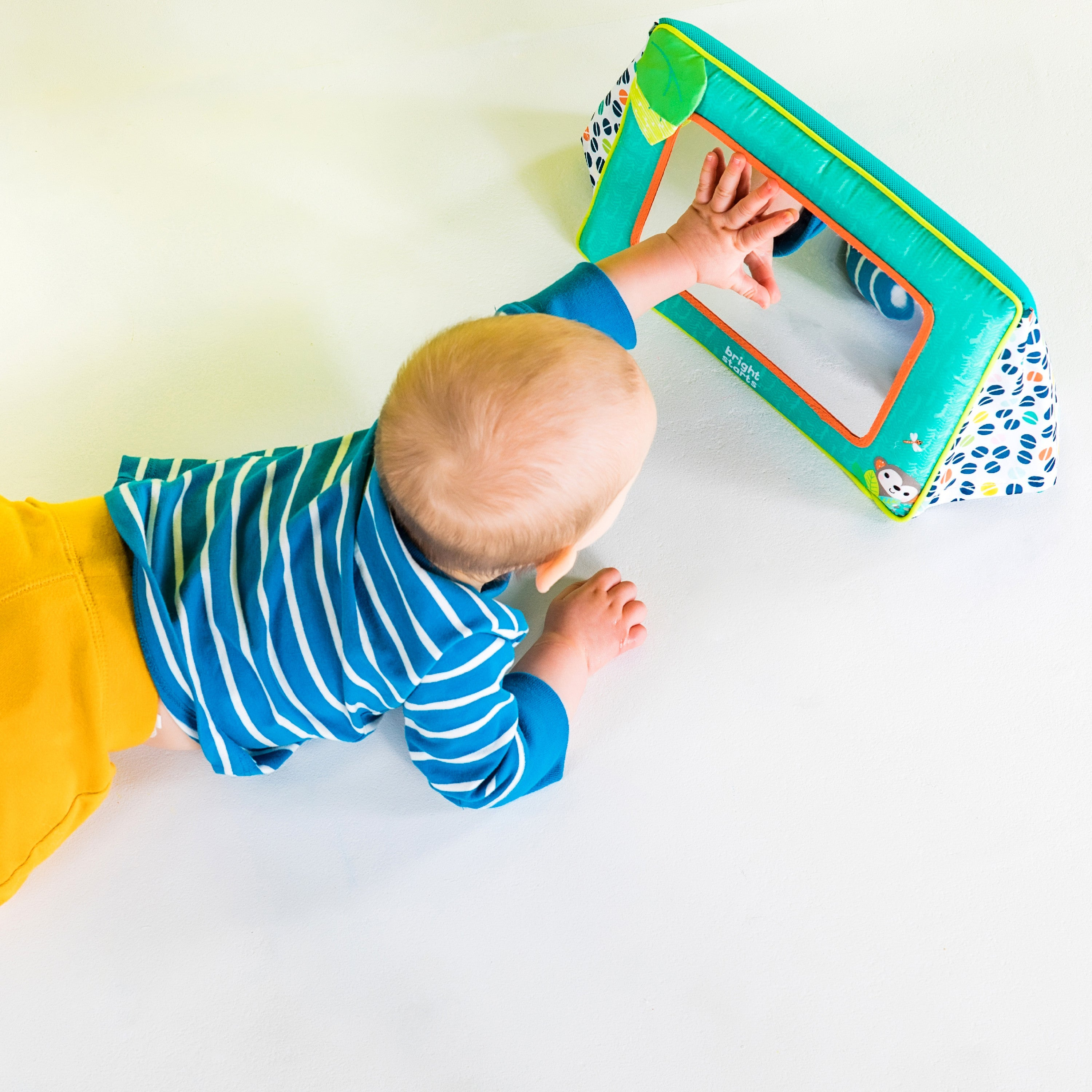 Ein Baby greift nach einem Spielzeugspiegel. Das Baby trägt ein blau-weiß gestreiftes Hemd und gelbe Hose.