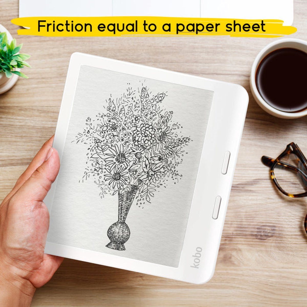 Una mano sostiene un lector Kobo con un boceto de flores en un jarrón. Café, gafas y planta sobre una mesa de madera.